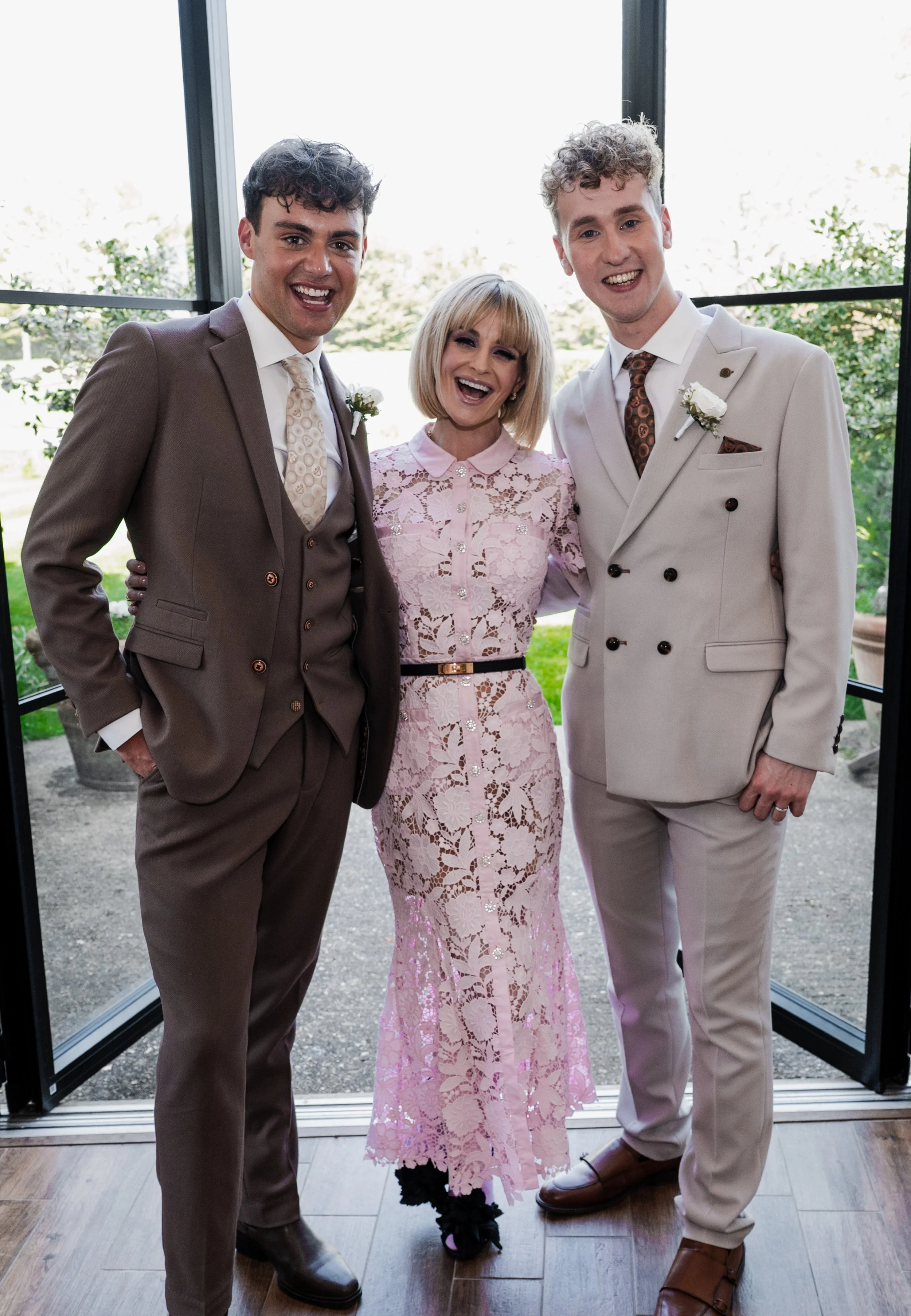 Two grooms in weddding attire stood either side of a smaller blonde woman wearing a pink dress