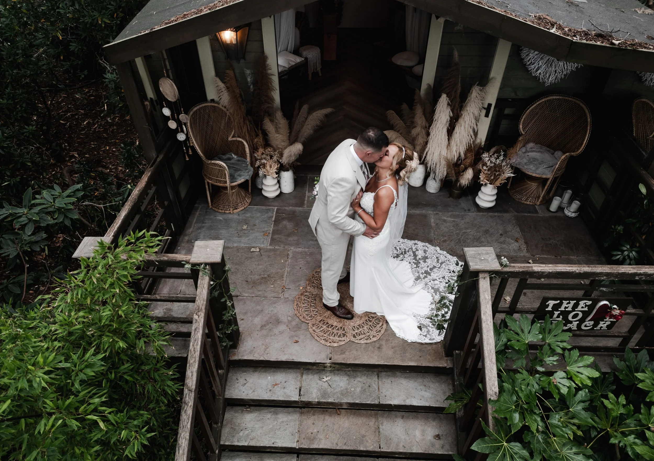 The Love Shack - Drone image of groom and bride in wedding attire, stood on top of a wooden staircase.
