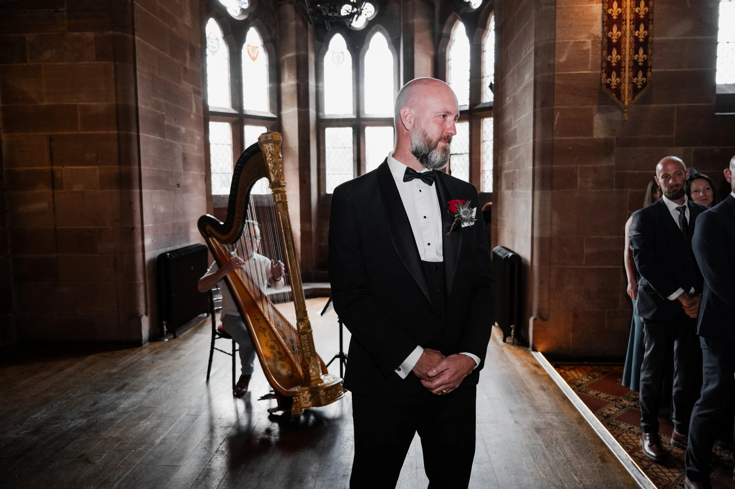 Peckforton Castle - Groom in wedding attire waiting at the top of the aisle with a harpist behind him 