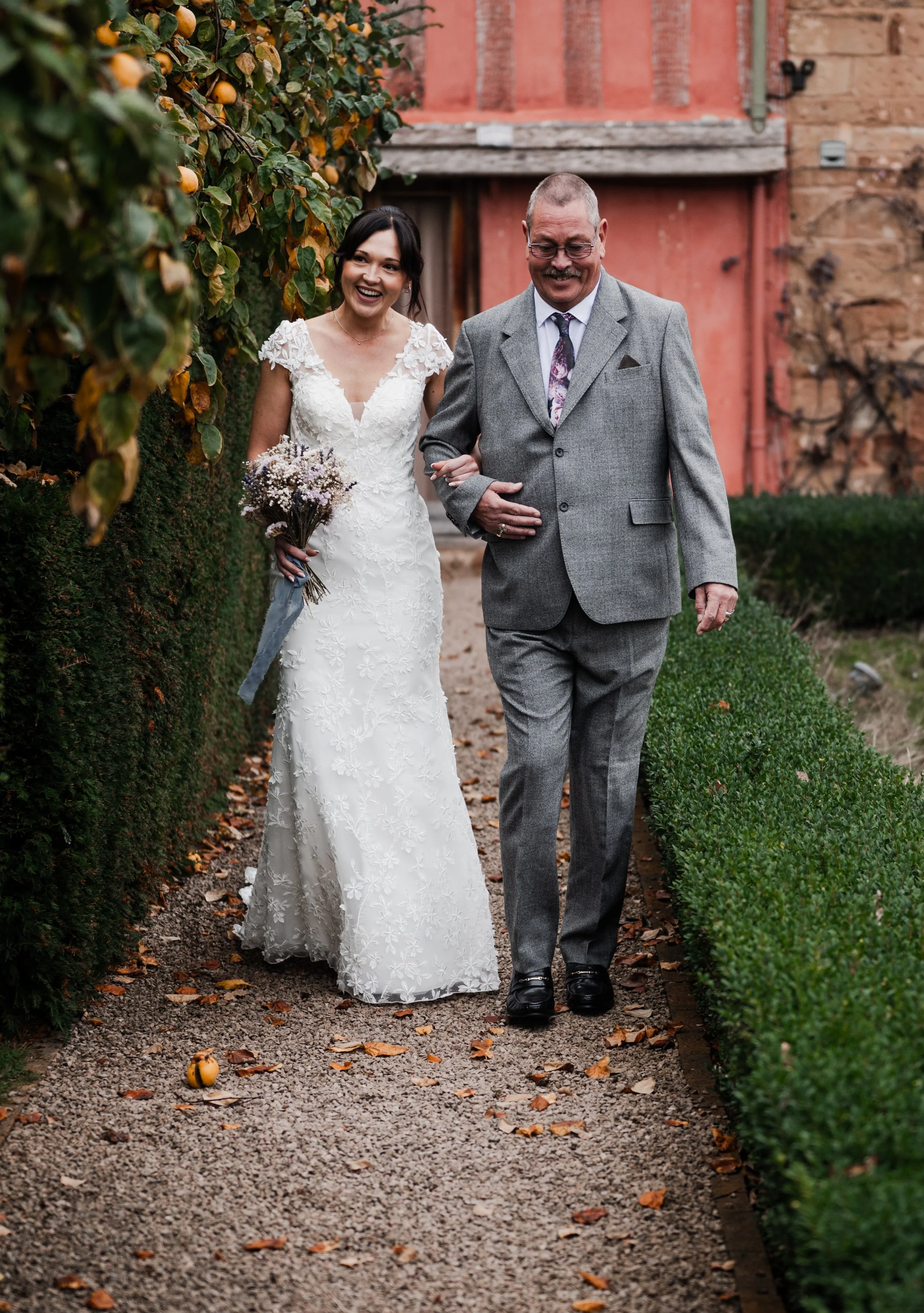 A bride in a white lace wedding dress walking arm-in-arm with a man in a gray suit on a garden path with bushes and fallen leaves pauntley court, Gareth roy photography