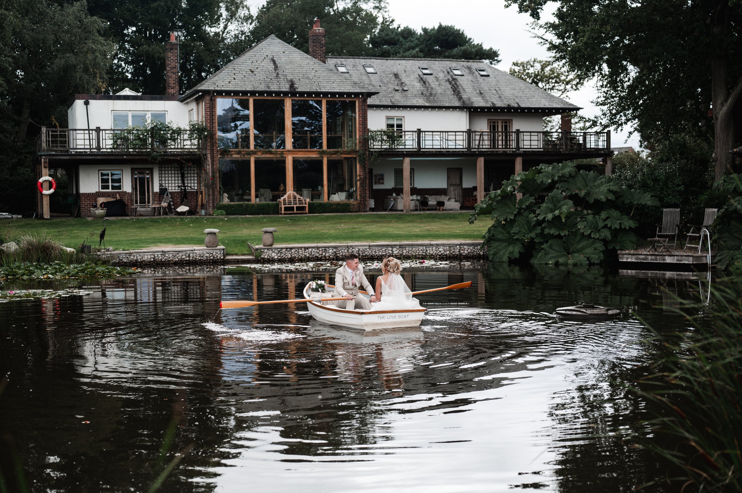 The Love Shack - Groom and bride in wedding attire, in a wooden boat in front of a large wedding venue