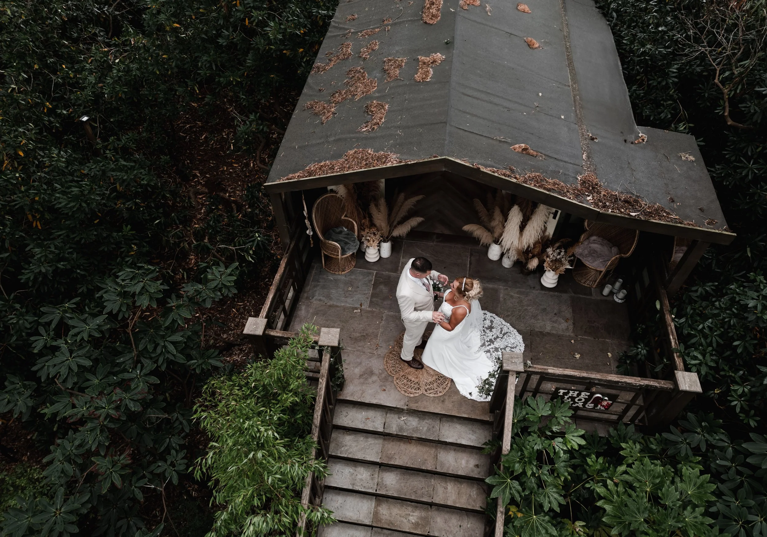 The Love Shack - Drone image of groom and bride in wedding attire, stood on top of a wooden staircase.