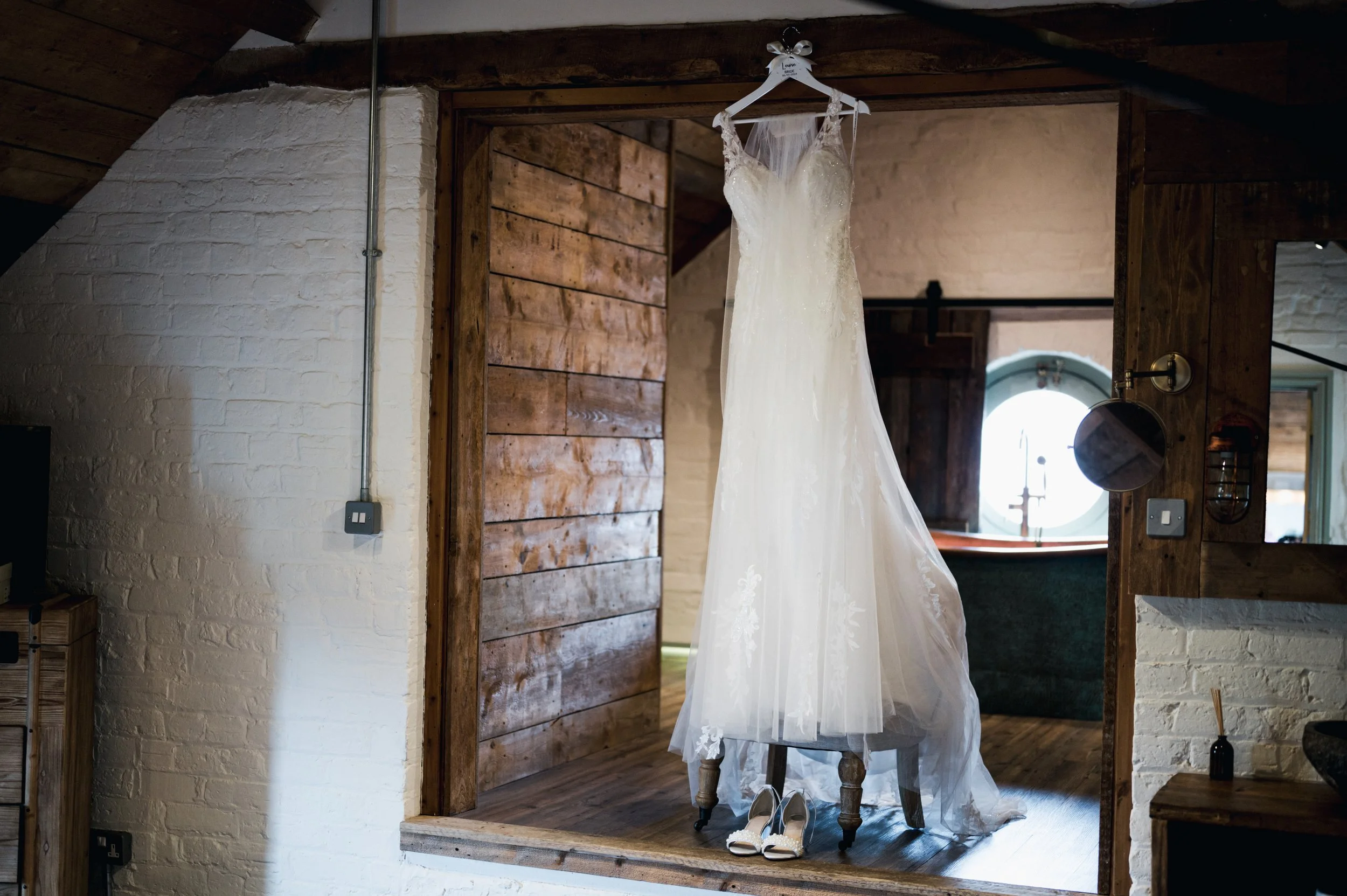 Wedding dress hanging on a hanger with a pair of white shoes below, displayed in a rustic room with wooden walls and a window in the background grange barn Whitchurch, Gareth roy photography