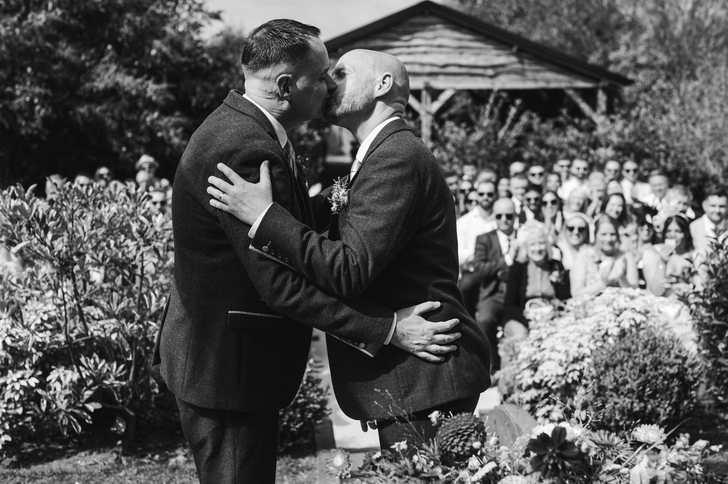 Wedding photography Cheshire - Black and white image of two grooms kissing at the end of a wedding ceremony in front of wedding guests 
