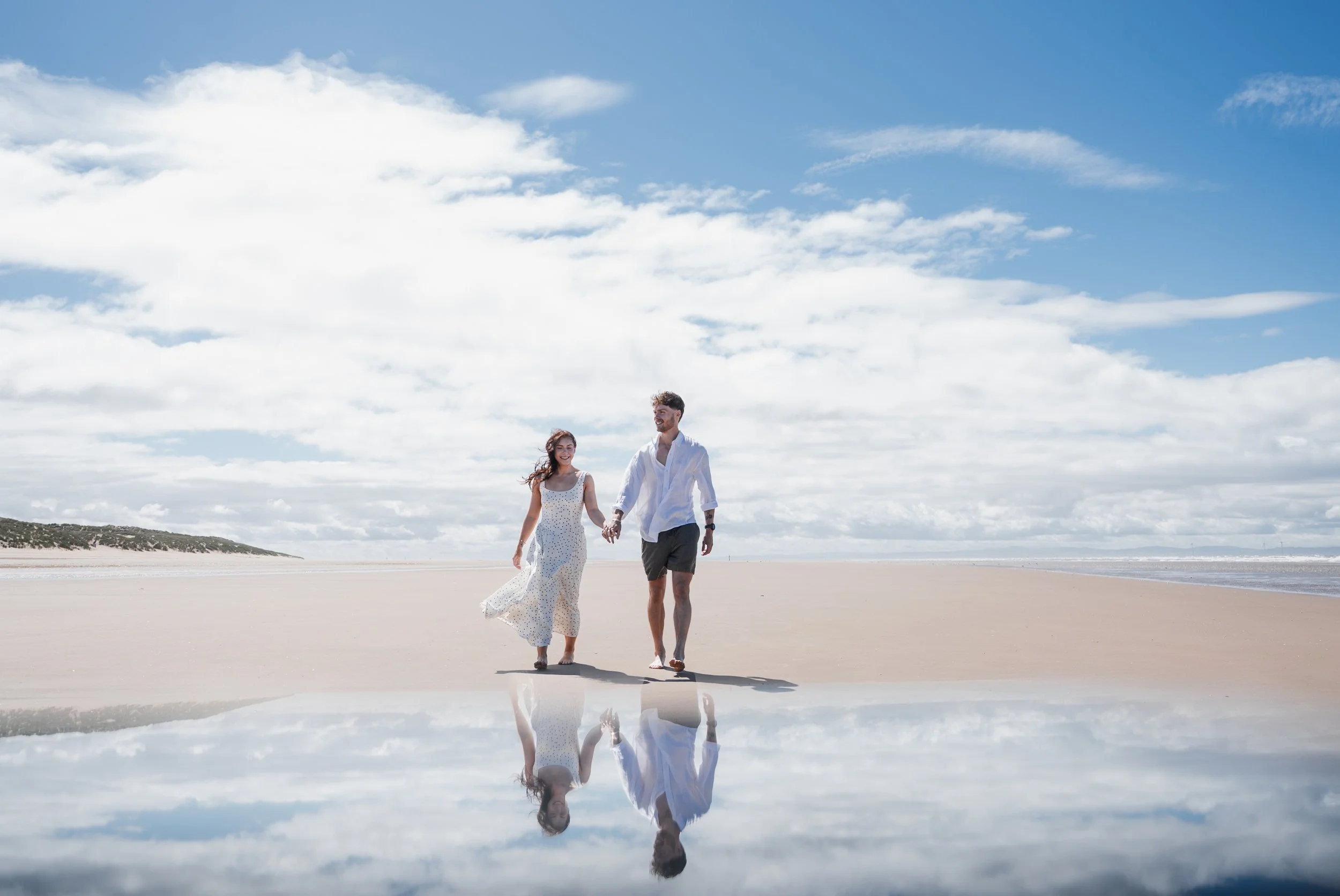 Formby Beach Engagement - Man and woman wearing white and black clothes on a beach , holding each other's hands and smiling while image is reflected up in the water 