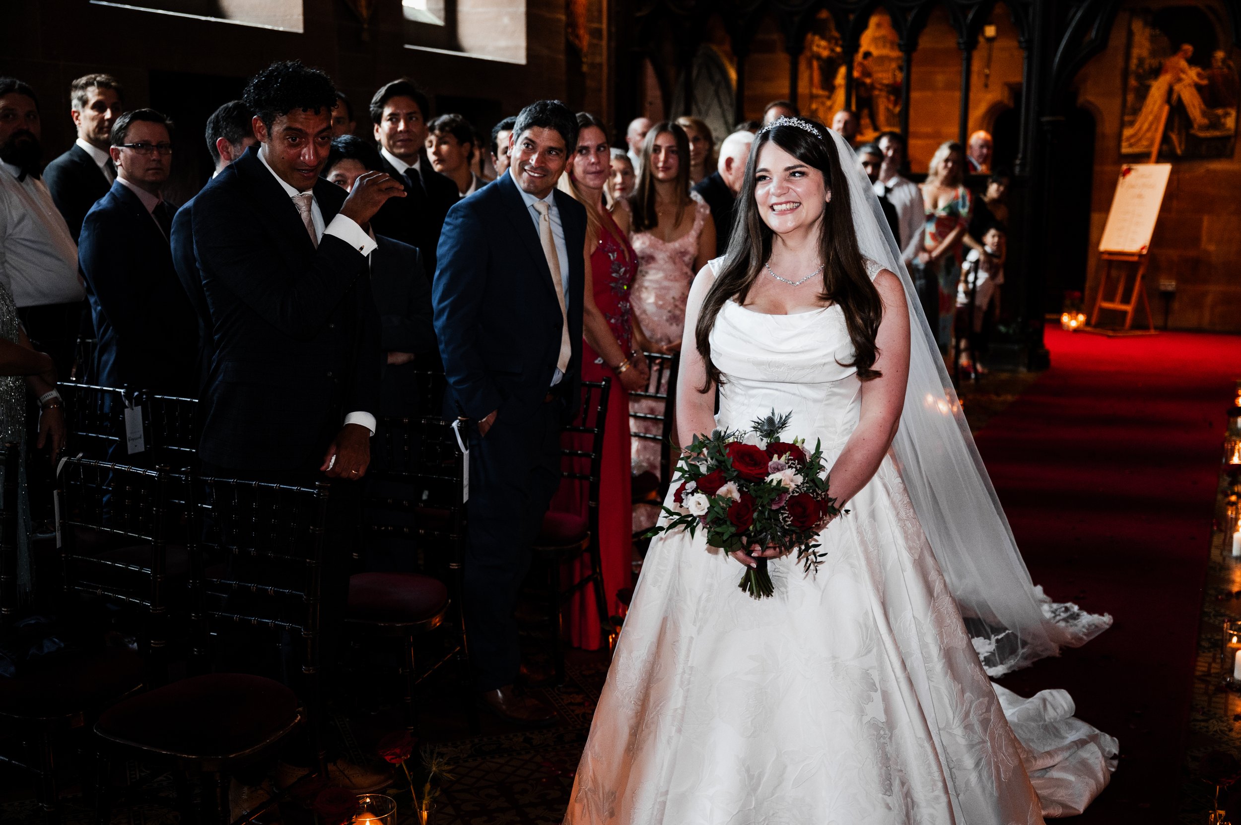 Peckforton Castle - a bride in wedding attire walking down the aisle surrounded by smiling wedding guests