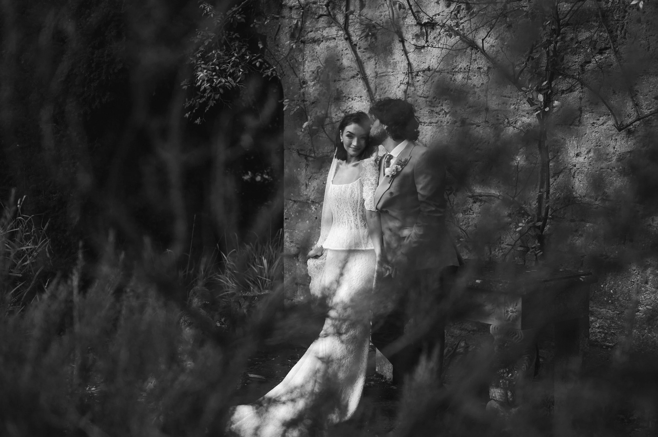 Black and white photo of a woman in a white lace wedding dress and a man in a suit standing close together against a stone wall, partially obscured by blurred tree branches in the foreground at sudeley castle, Gareth roy photography