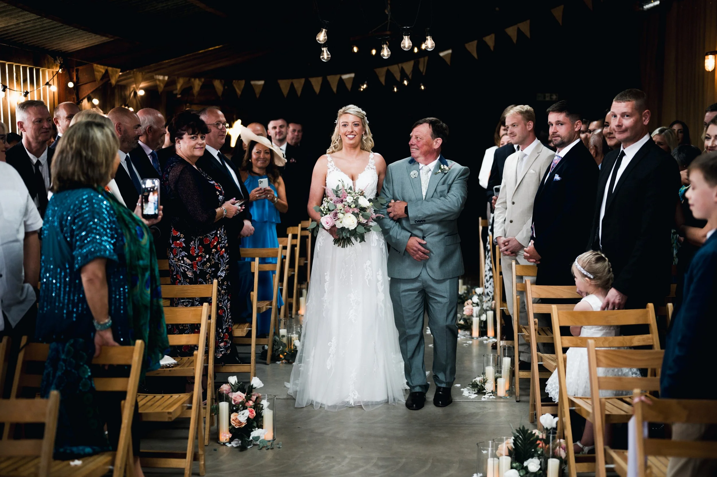Bride in a white gown walking down the aisle on her wedding day, accompanied by a man in a light gray suit, with guests standing on both sides watching and taking photos grange barn Whitchurch, Gareth roy photography