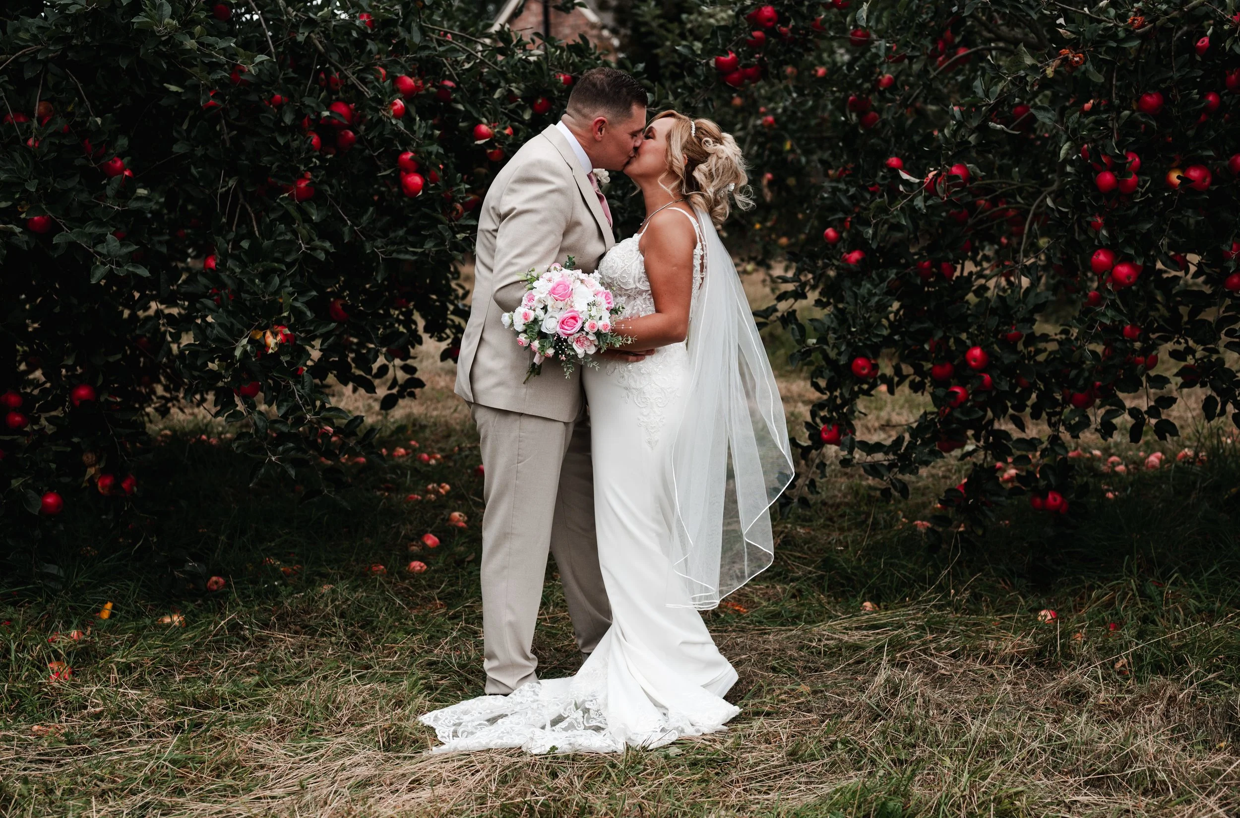 The Love Shack - groom and bride in wedding attire, kissing in front of foliage with red and pink flowers. 