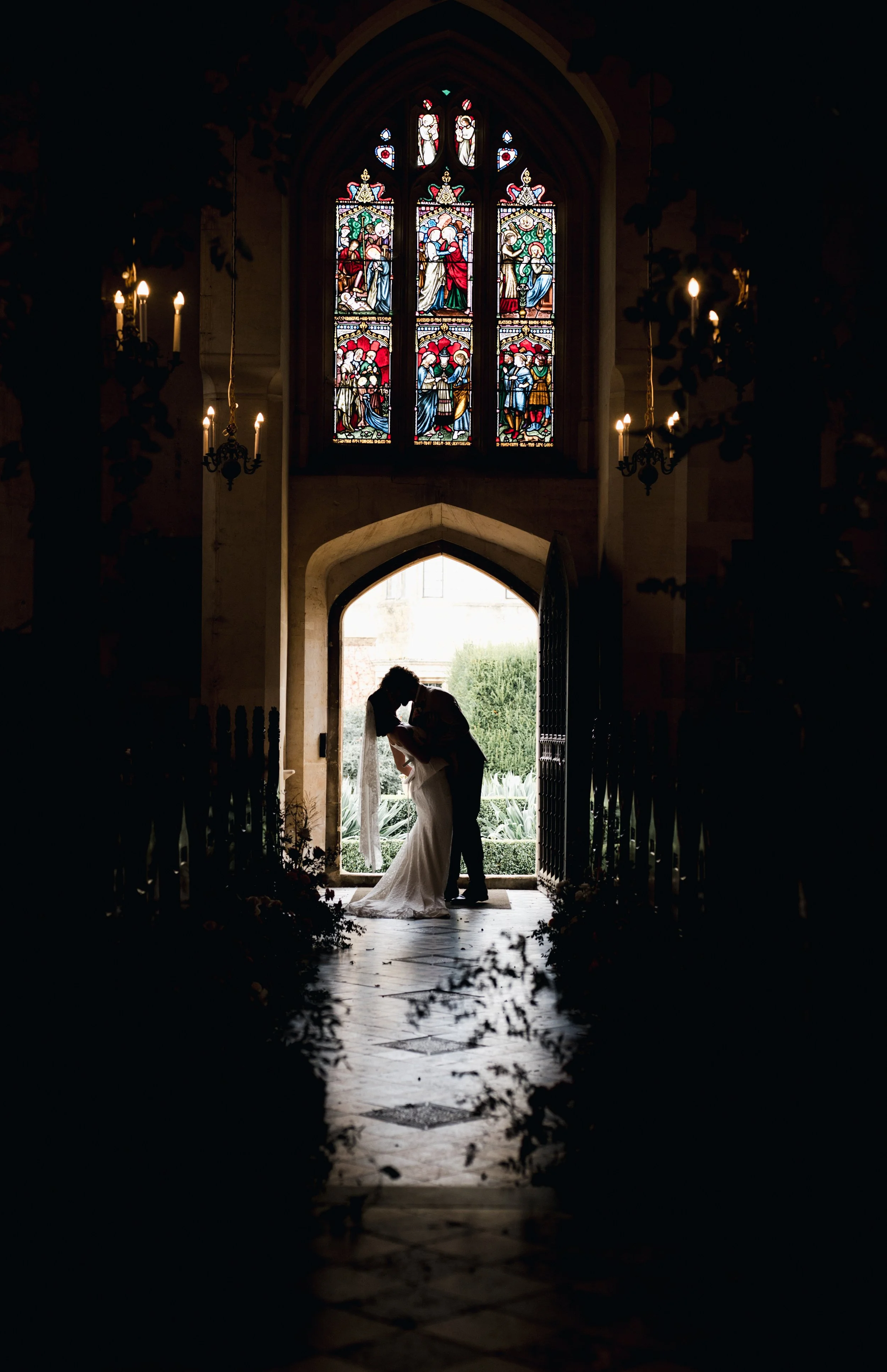 Silhouette of a bride and groom sharing a kiss inside a church with stained glass windows and open doors, with natural light filtering in from outside - Sudeley Castle