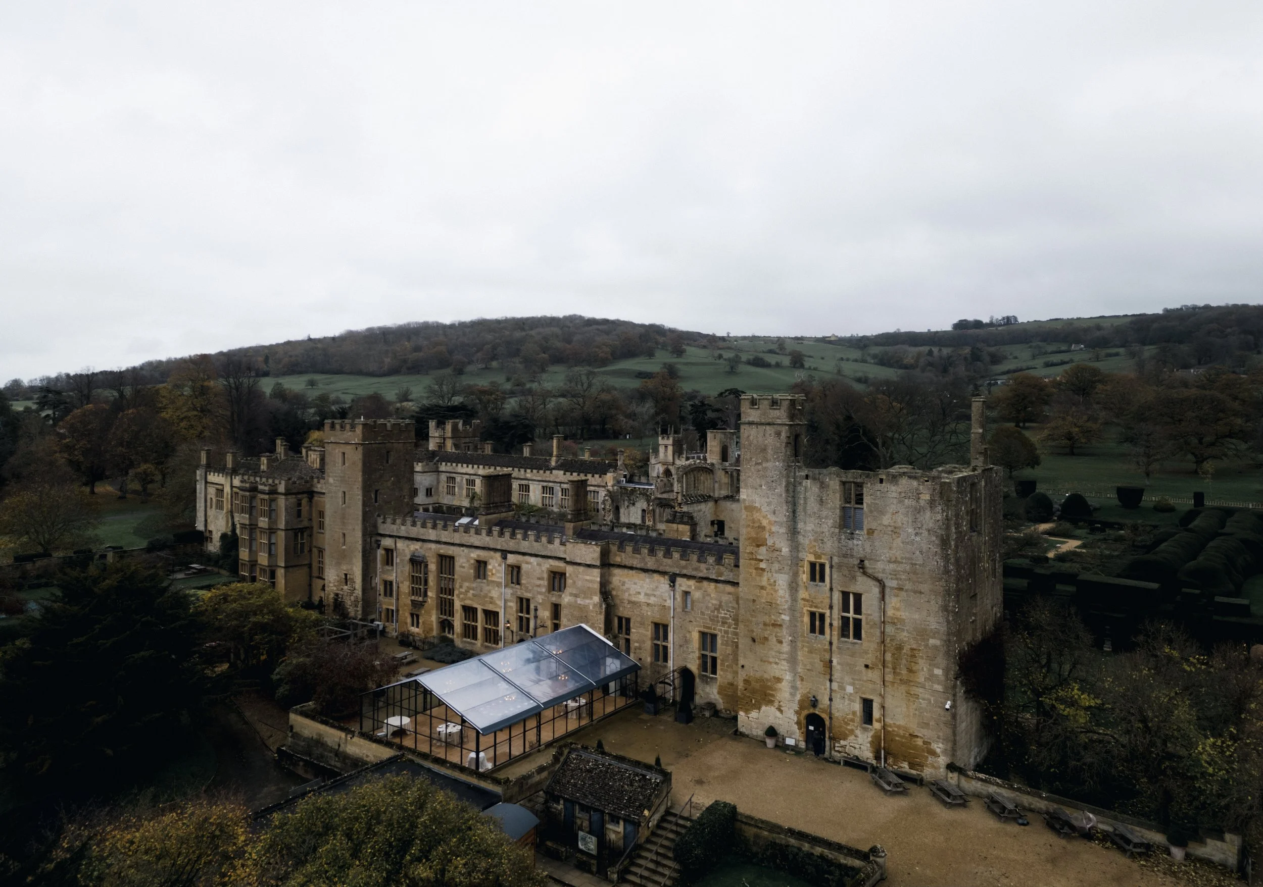 A large historic castle with stone walls, towers, and battlements, surrounded by trees and open landscape under an overcast sky. at sudeley castle, Gareth roy photography