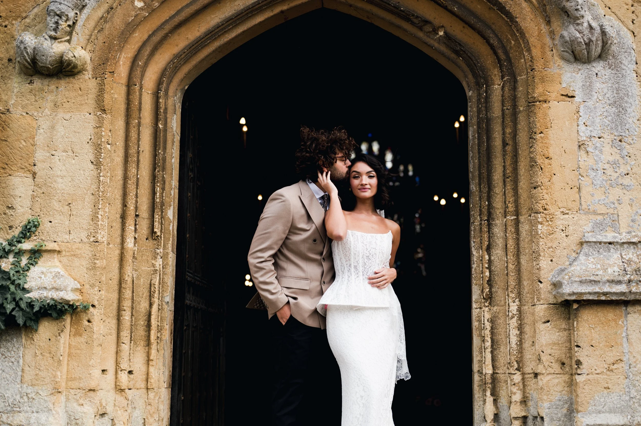 A newlywed couple standing in a stone archway, with the groom whispering to the bride who is smiling, and the background appears dark with blurred lights at sudeley castle, Gareth roy photography
