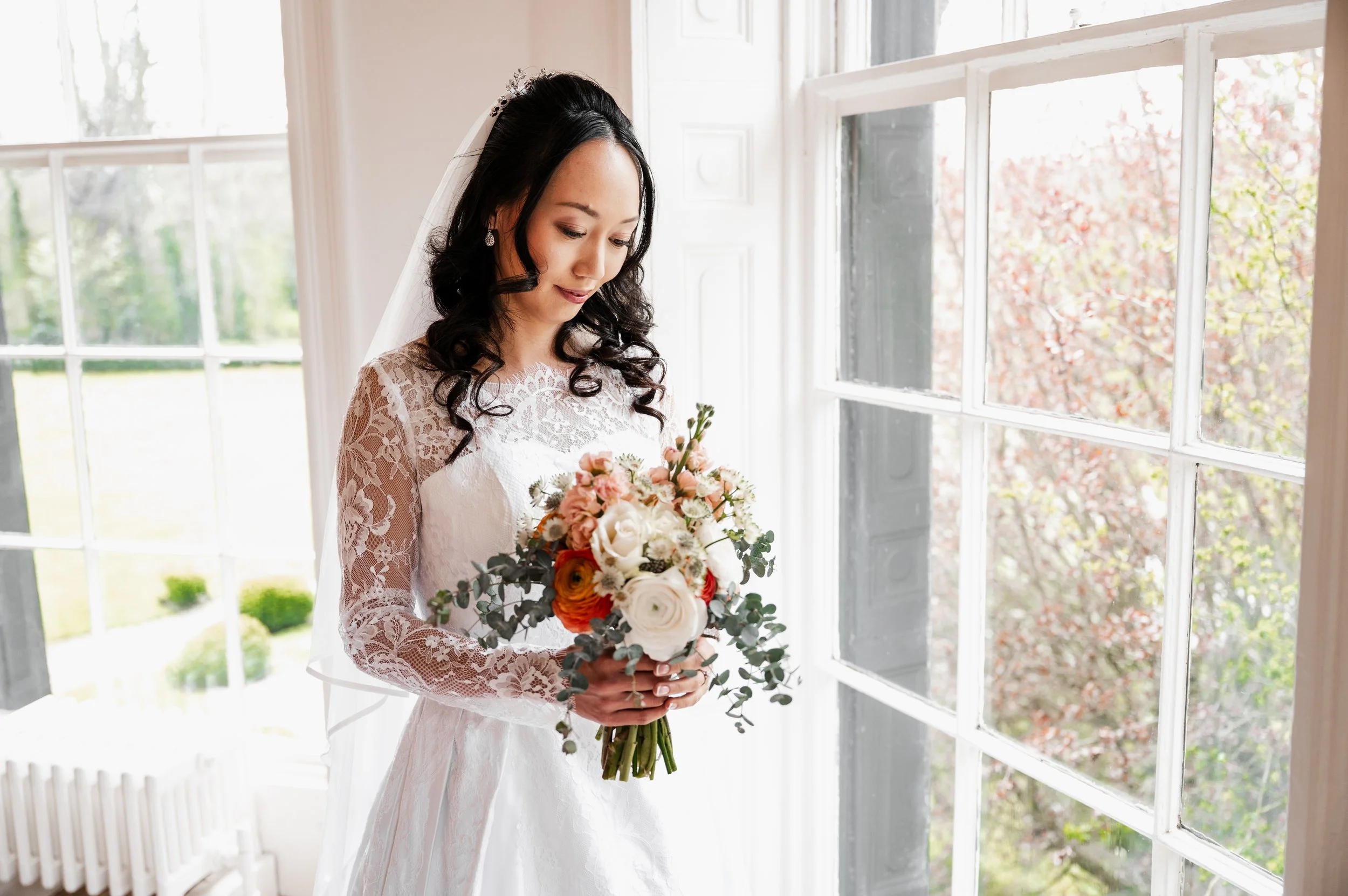 Wedding Photographer Cheshire - Bride in a lace dress stood in a light, airy window bay and looking at her bouquet of colourful flowers