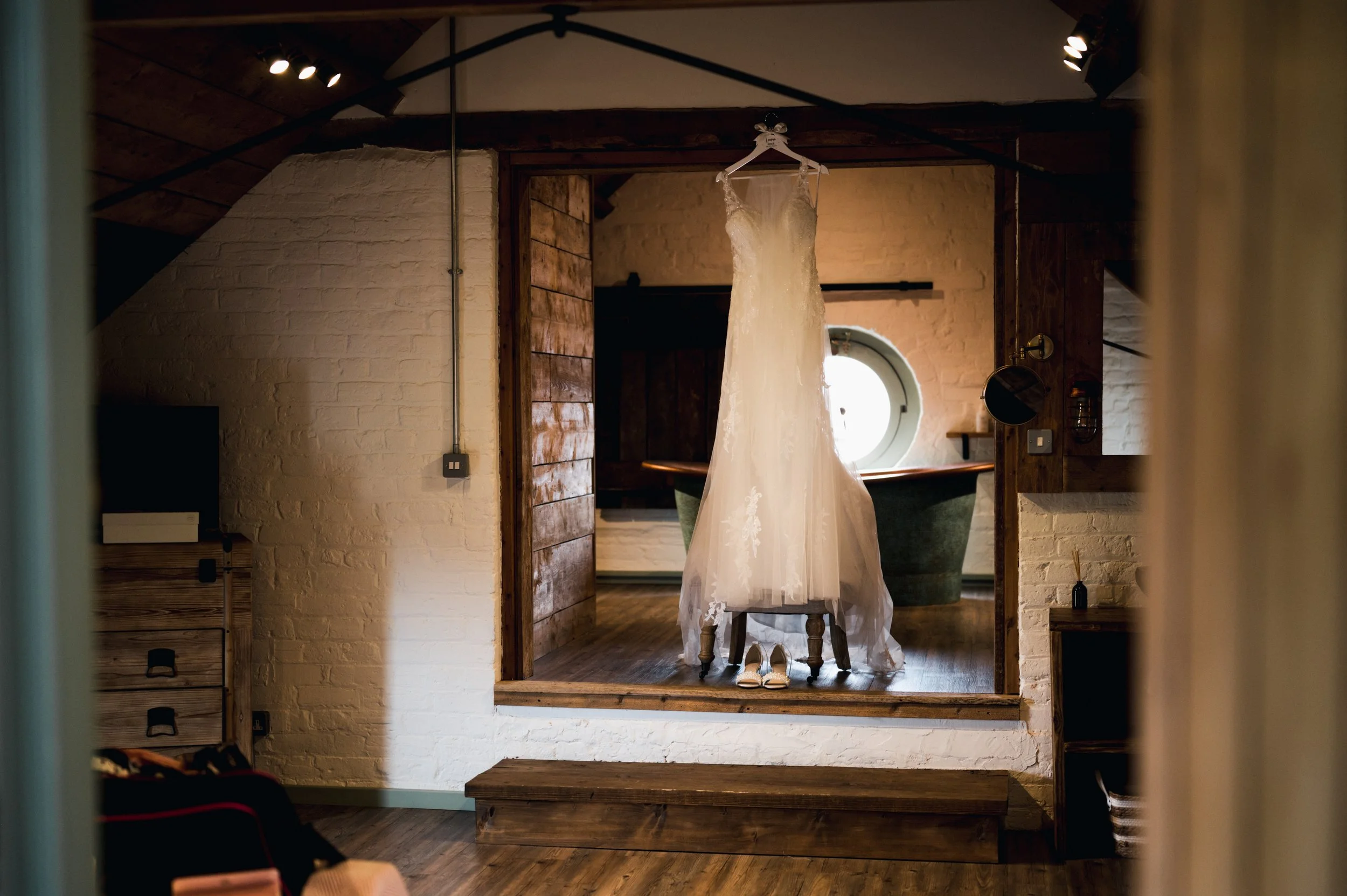 Wedding dress hanging on a hanger in a rustic room with white brick walls and wooden accents, overlooking a bathtub and a round window in the background grange barn Whitchurch, Gareth roy photography