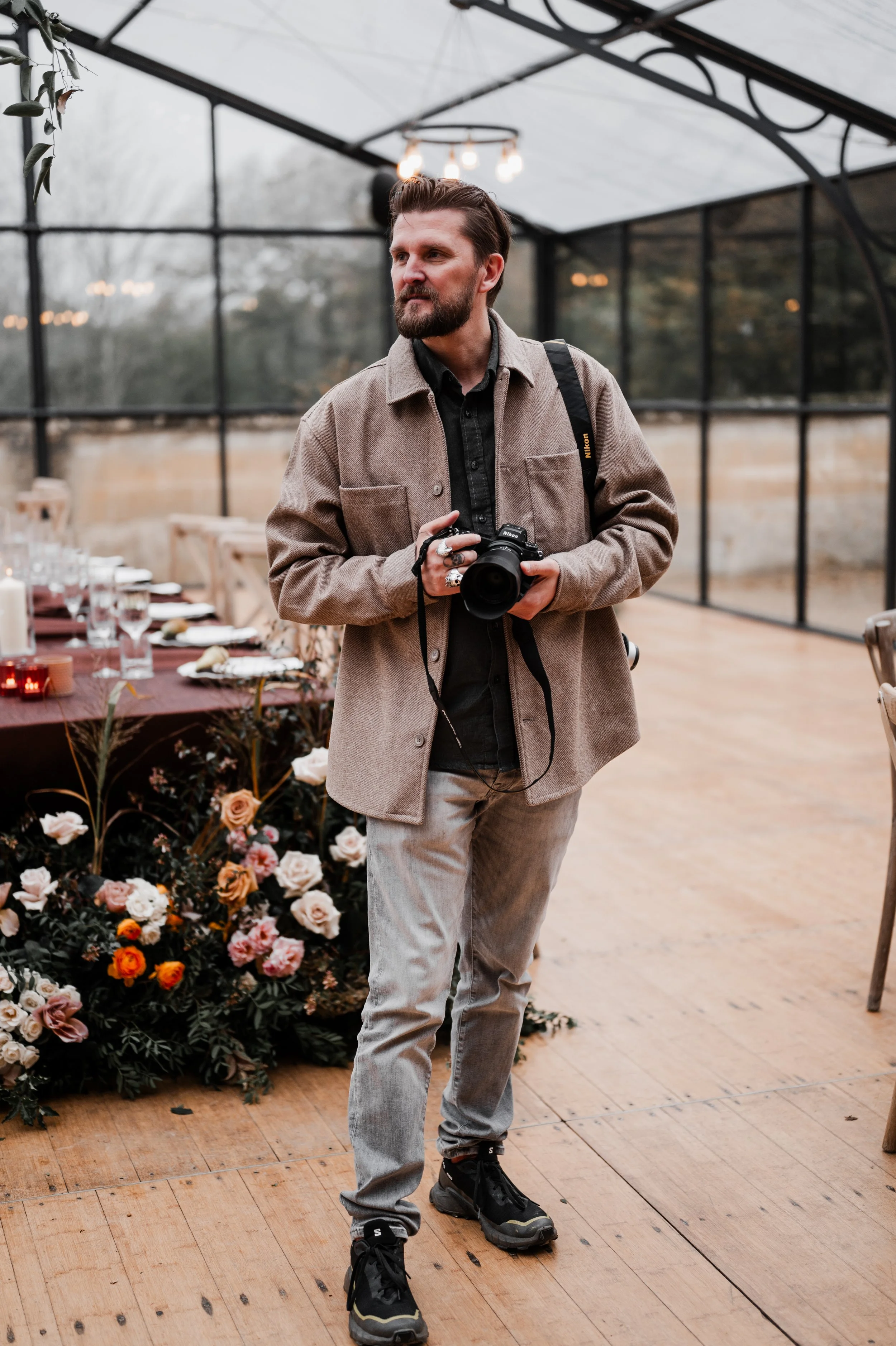 A man with brown hair and a beard holding a camera stands in a greenhouse or glass-walled structure with flowers and table settings in the background.