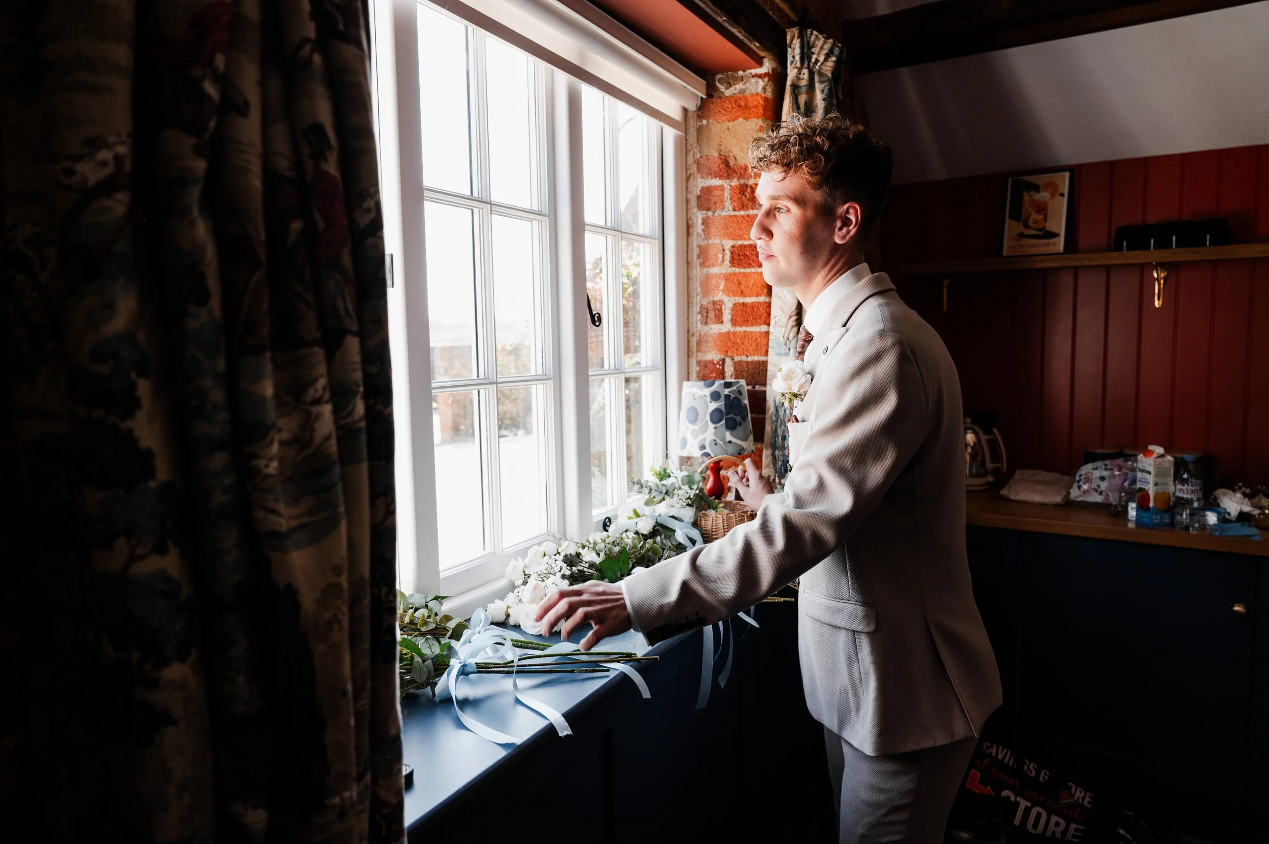 Groom in light brown suit looking out over gardens through a window