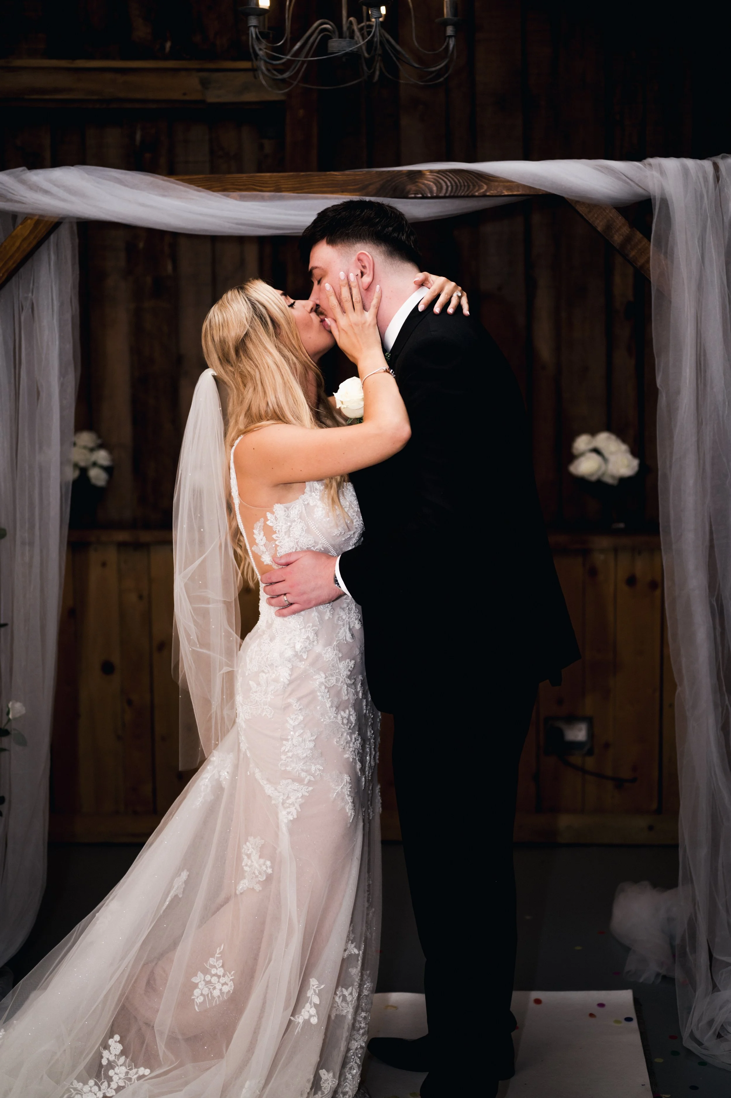 A bride and groom sharing a kiss under a wedding arch decorated with white tulle and flowers inside a rustic wooden venue.