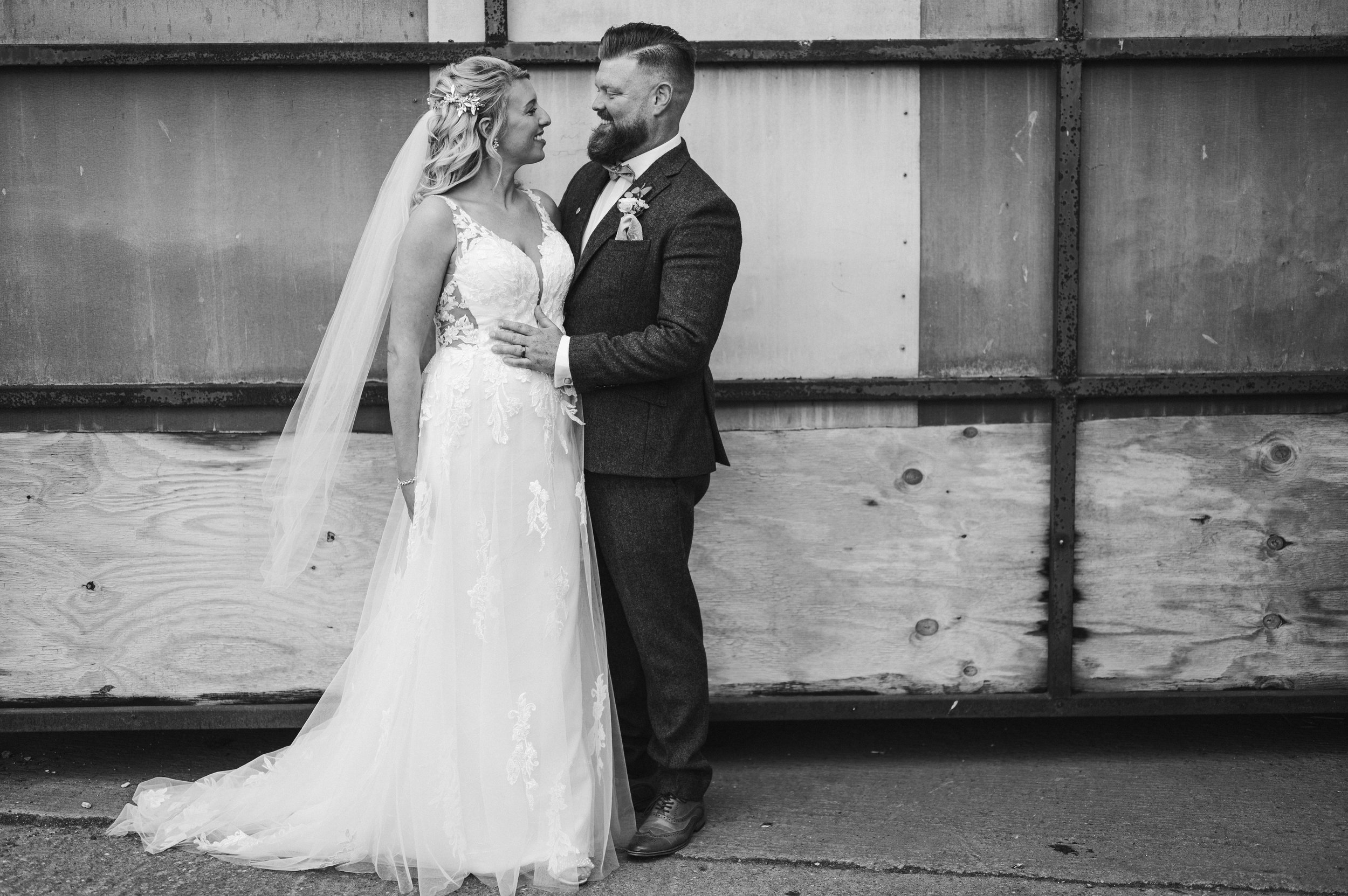 Black and white photo of a bride and groom standing close together, smiling at each other, against a rustic wooden wall background grange barn Whitchurch, Gareth roy photography