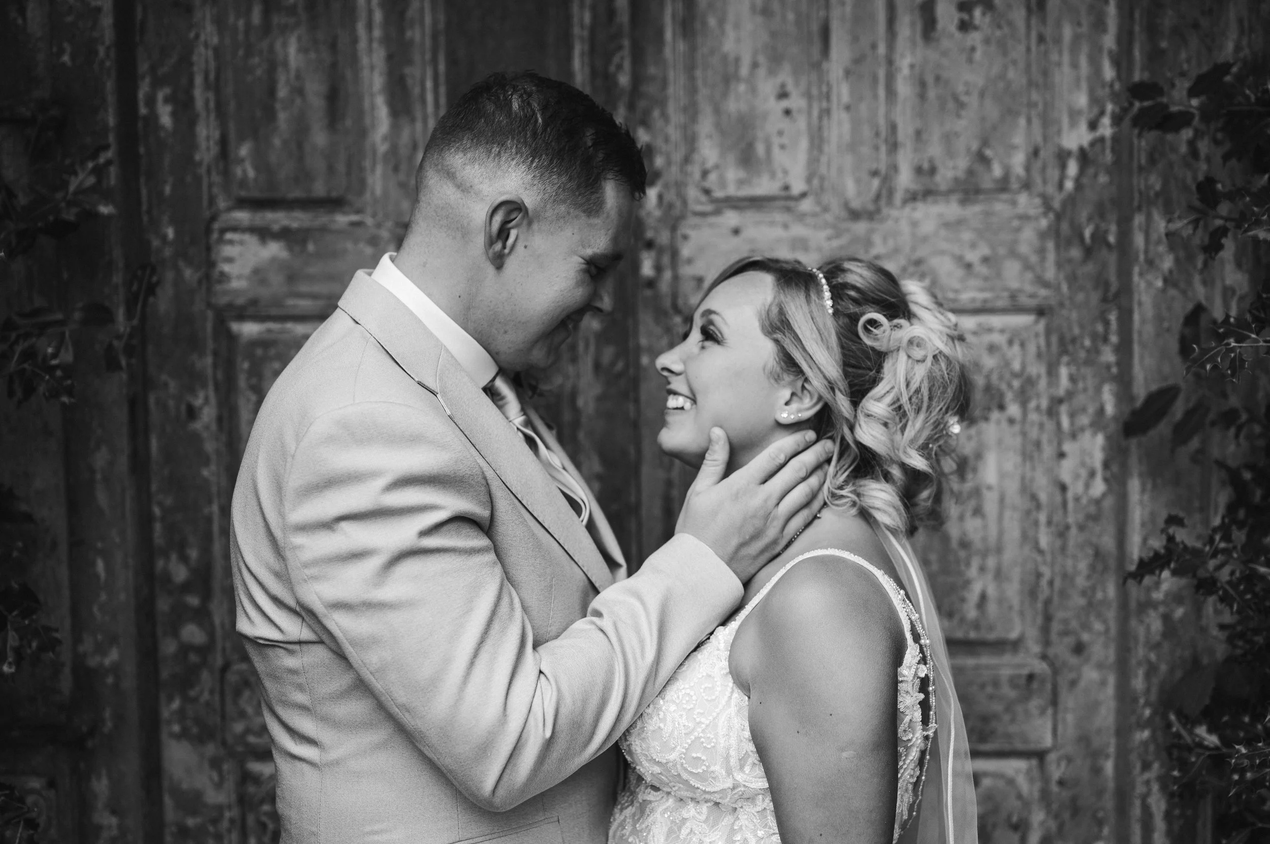 The Love Shack - black and white image of groom and bride in wedding attire, looking at each other and smiling in front of a wooden door. 