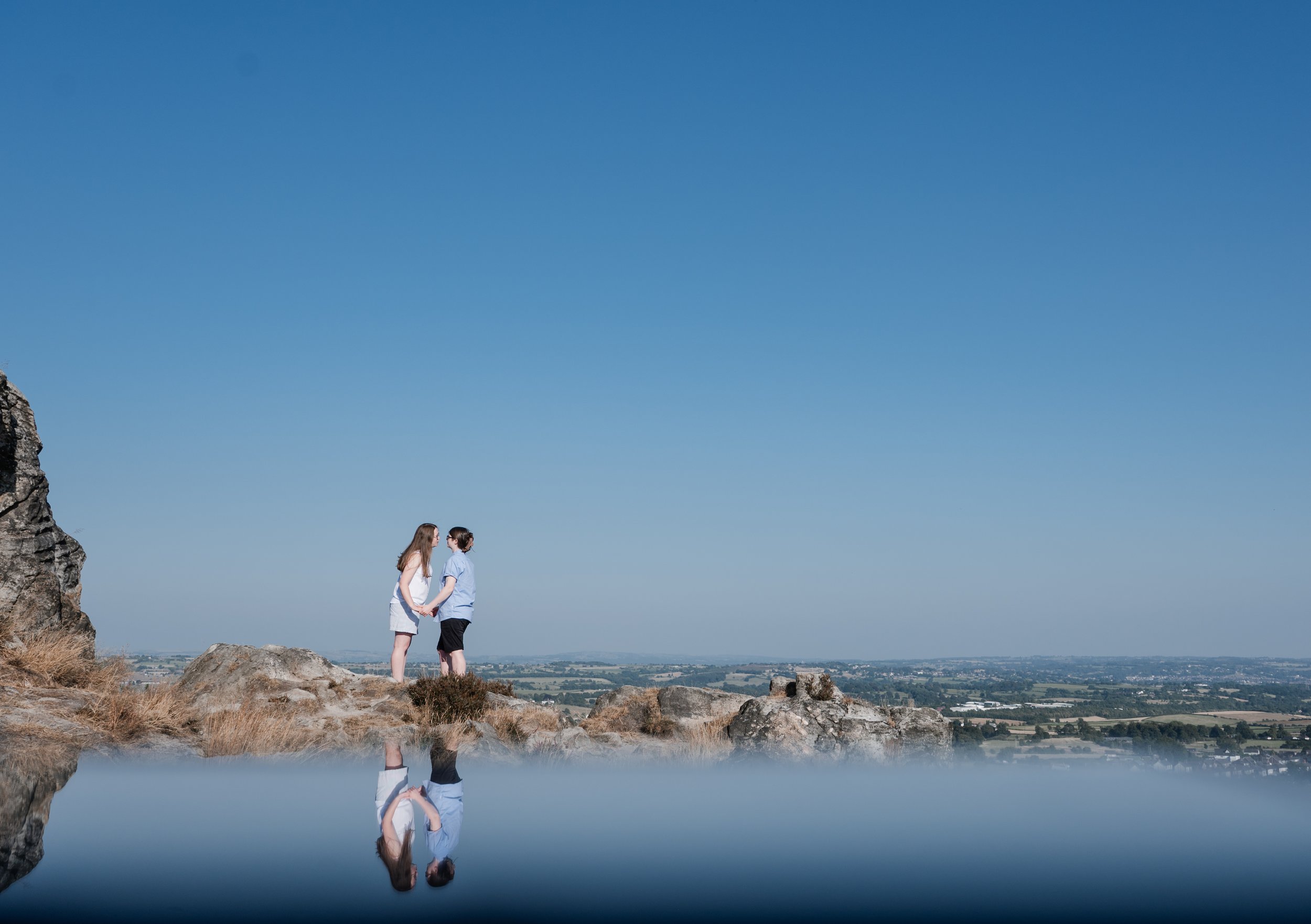 Mow Cop Engagement Shoot - Two women stood by a lake, holding hands and looking at each other with view of Cheshire behind them. 