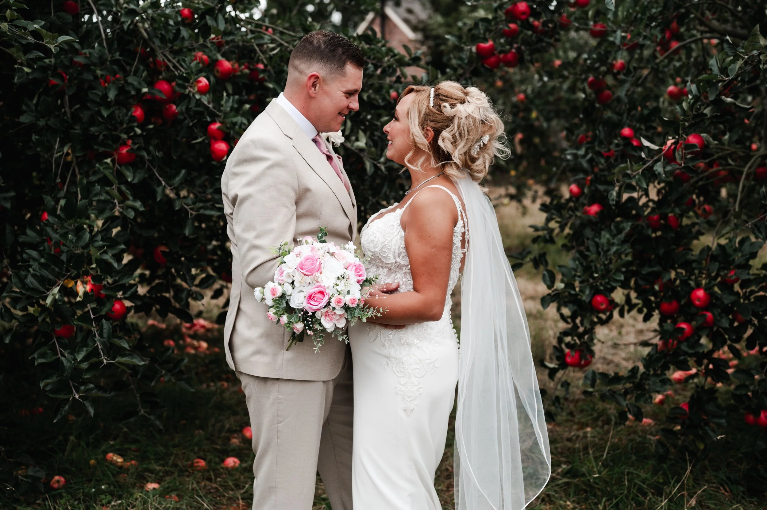 The Love Shack - groom and bride in wedding attire, looking at each other and smiling in front of foliage with red and pink flowers. 