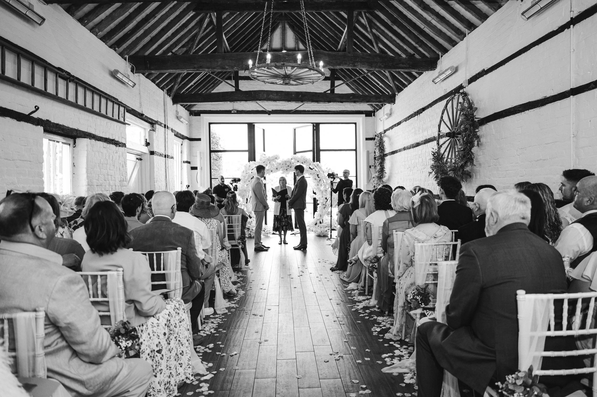 Black and white image of ceremony taking place from the bottom of the aisle with two grooms stood in front of large windows and a floral arch