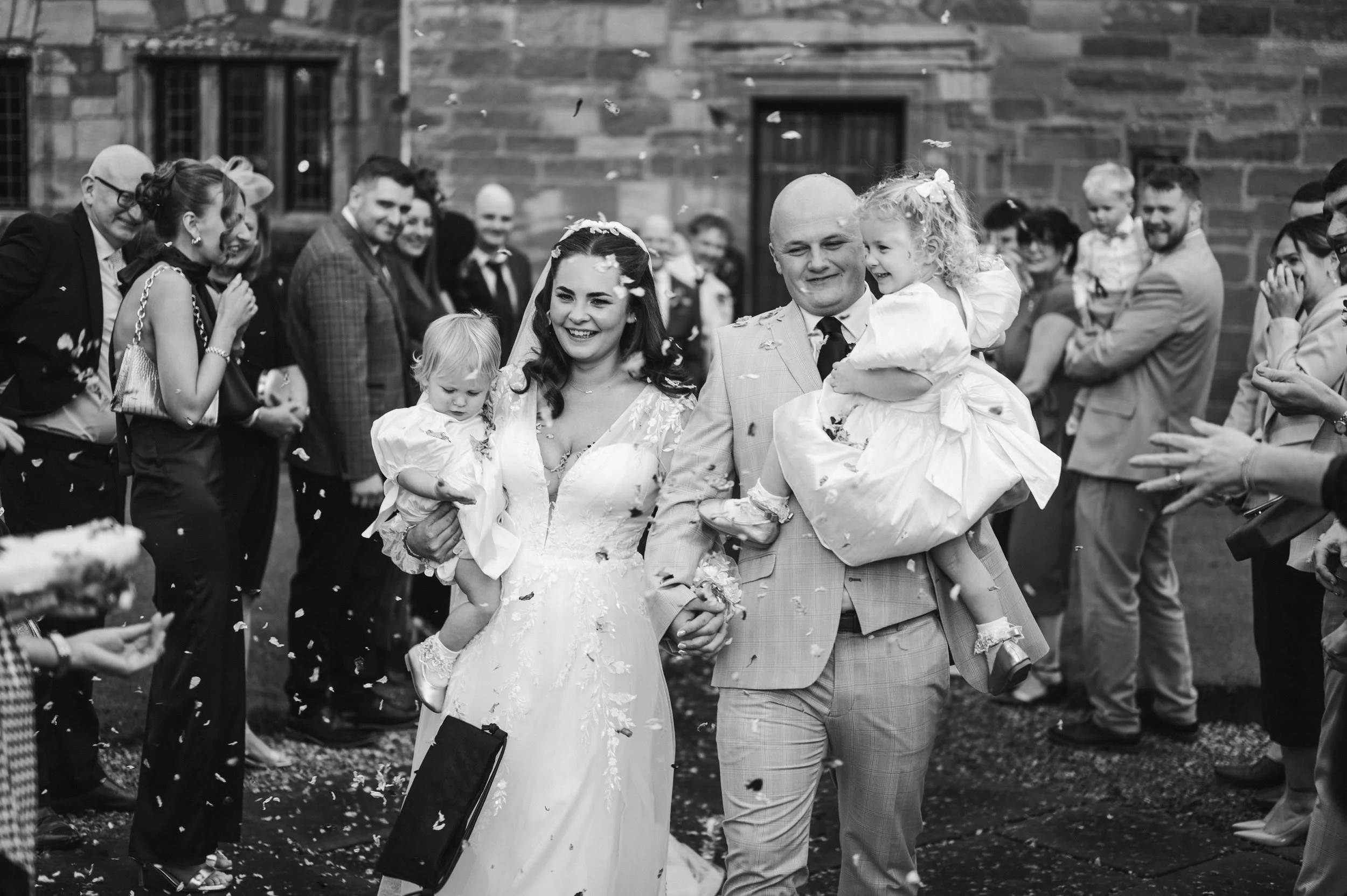 Cheshire Wedding Photographer - Bride and Groom in wedding attire holding small children while walking through wedding guests throwing confetti