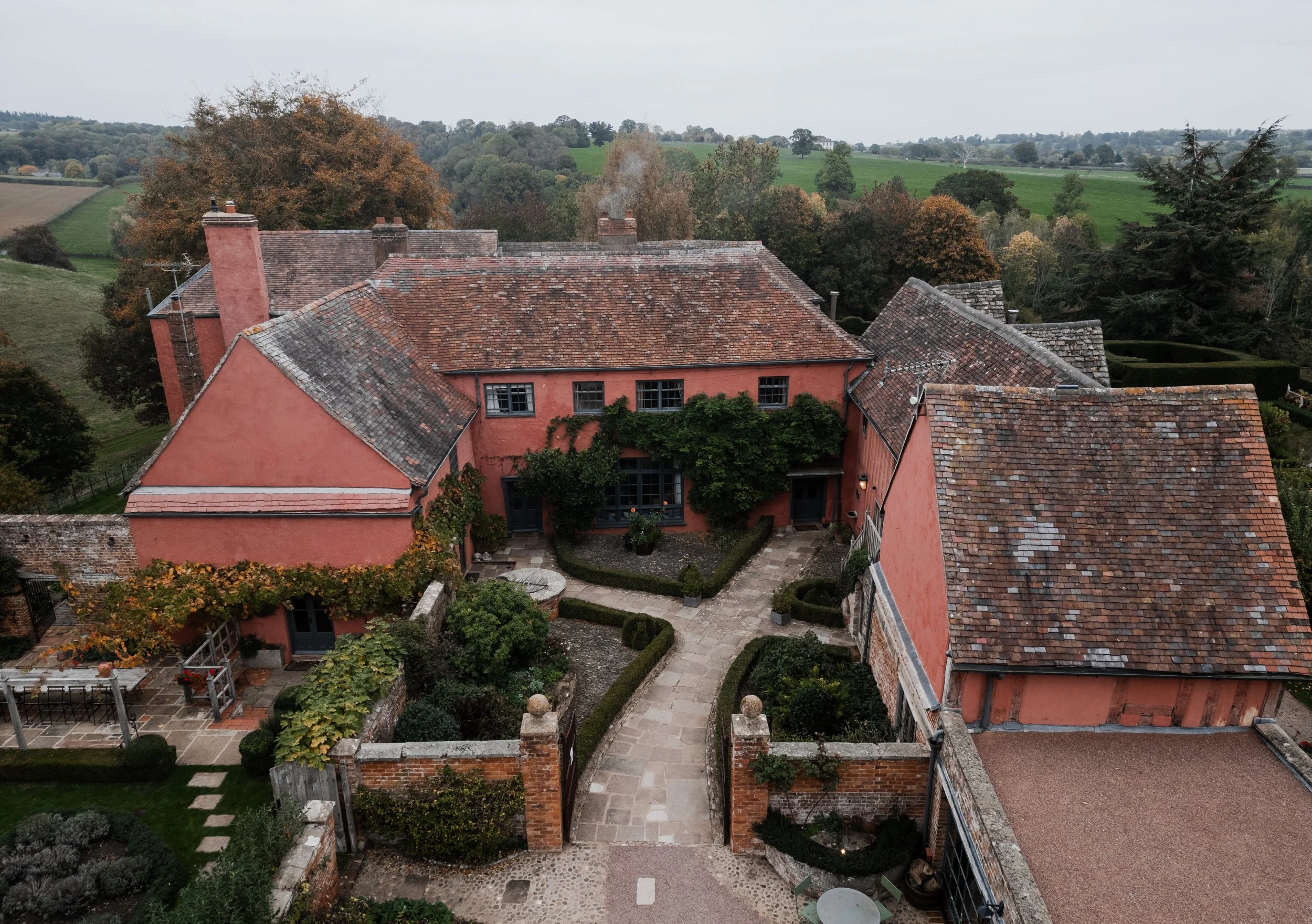 Aerial view of a large peach-colored country house with a courtyard, surrounded by landscaped gardens, trees, and countryside in the background at sudeley castle, Gareth roy photography