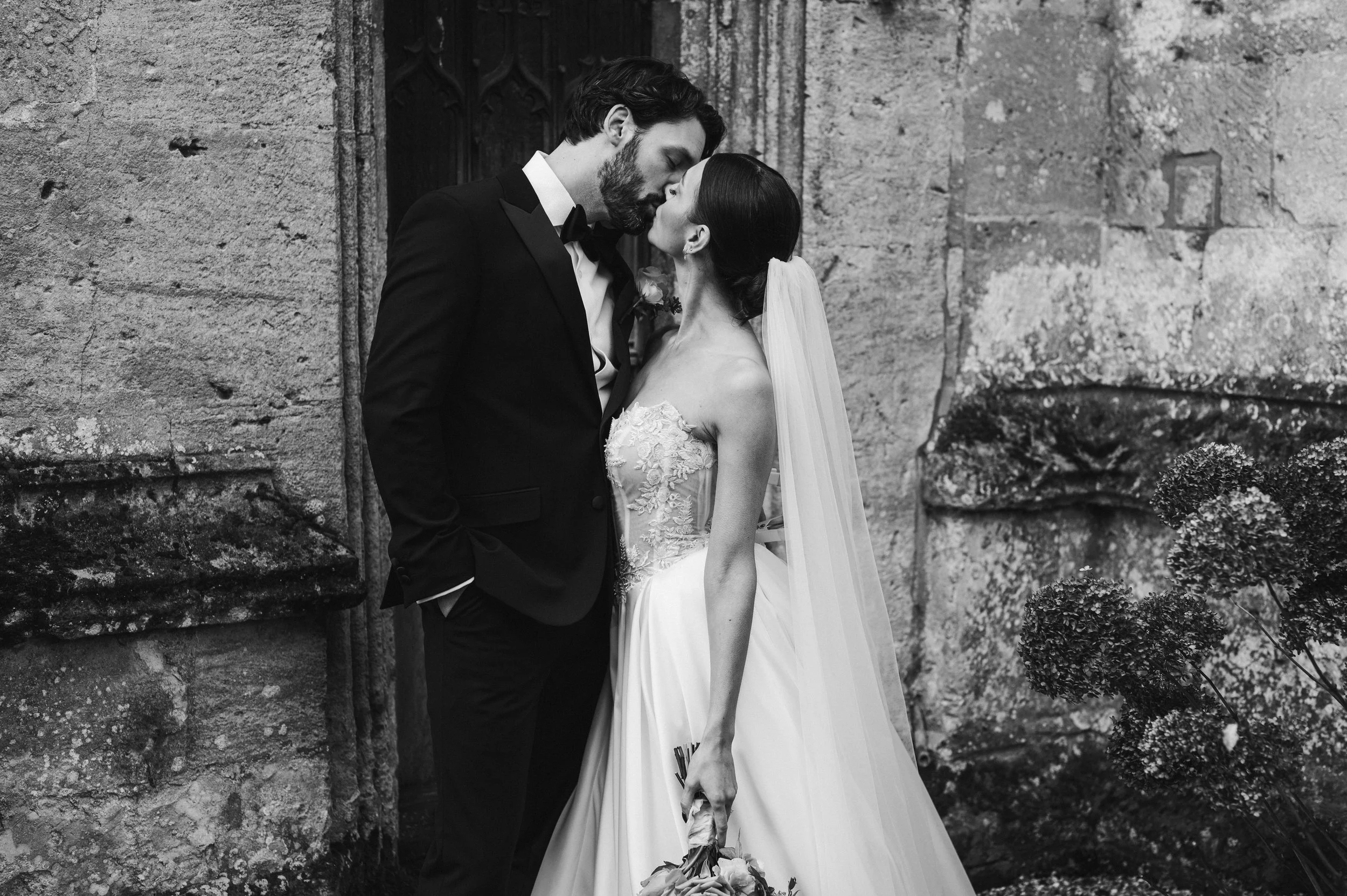 A black and white photo of a bride and groom kissing outside a stone building at sudeley castle, Gareth roy photography
