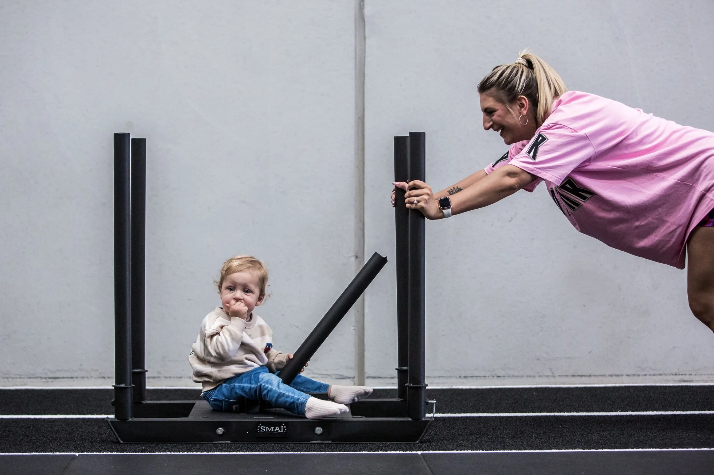A woman in a pink shirt pushing a small child sitting on a black exercise sled at Mumtribe Fitness in Carrum Downs