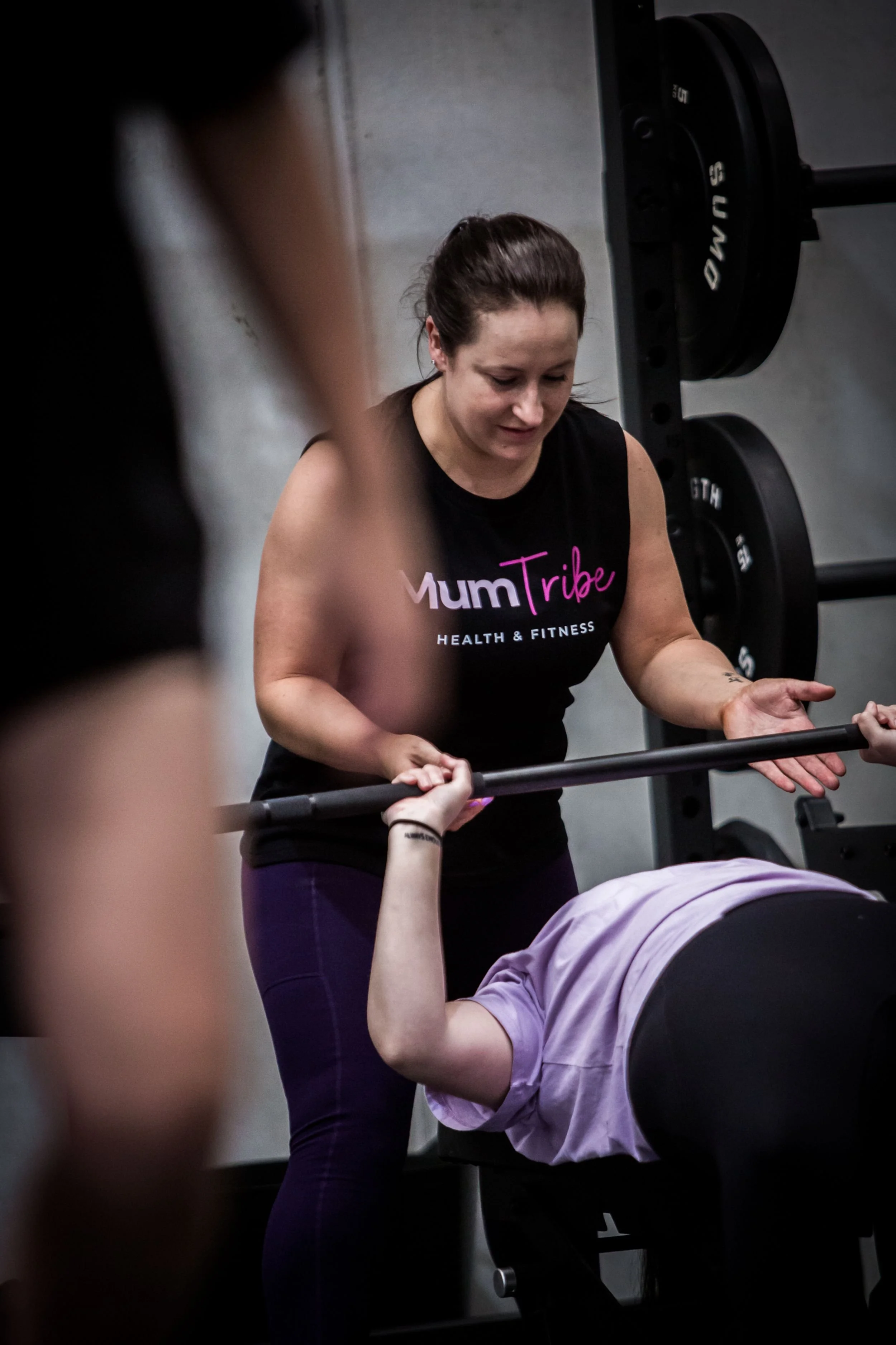 Head coach Kim in a black tank top with 'MumTribe' logo helps a person with a purple shirt do bench press at a gym.