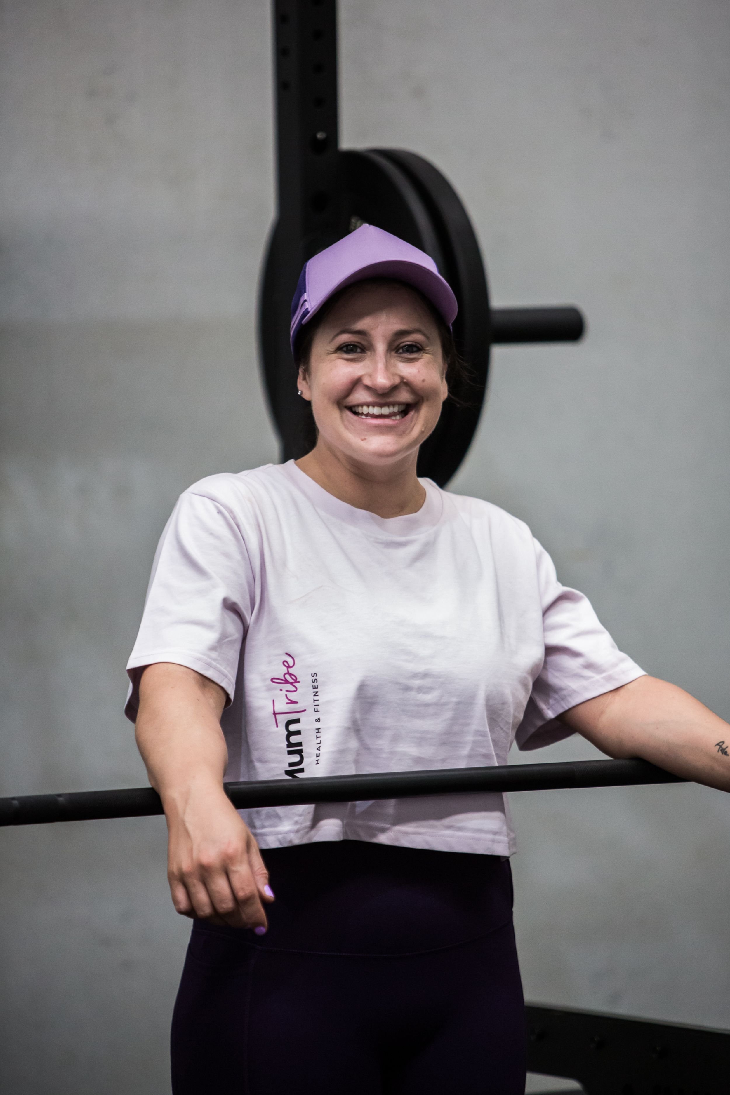 Kim, head coach at Mumtribe Fitness in Carrum Downs, smiling  and holding a barbell with a weight behind her, wearing a white T-shirt and a purple cap.