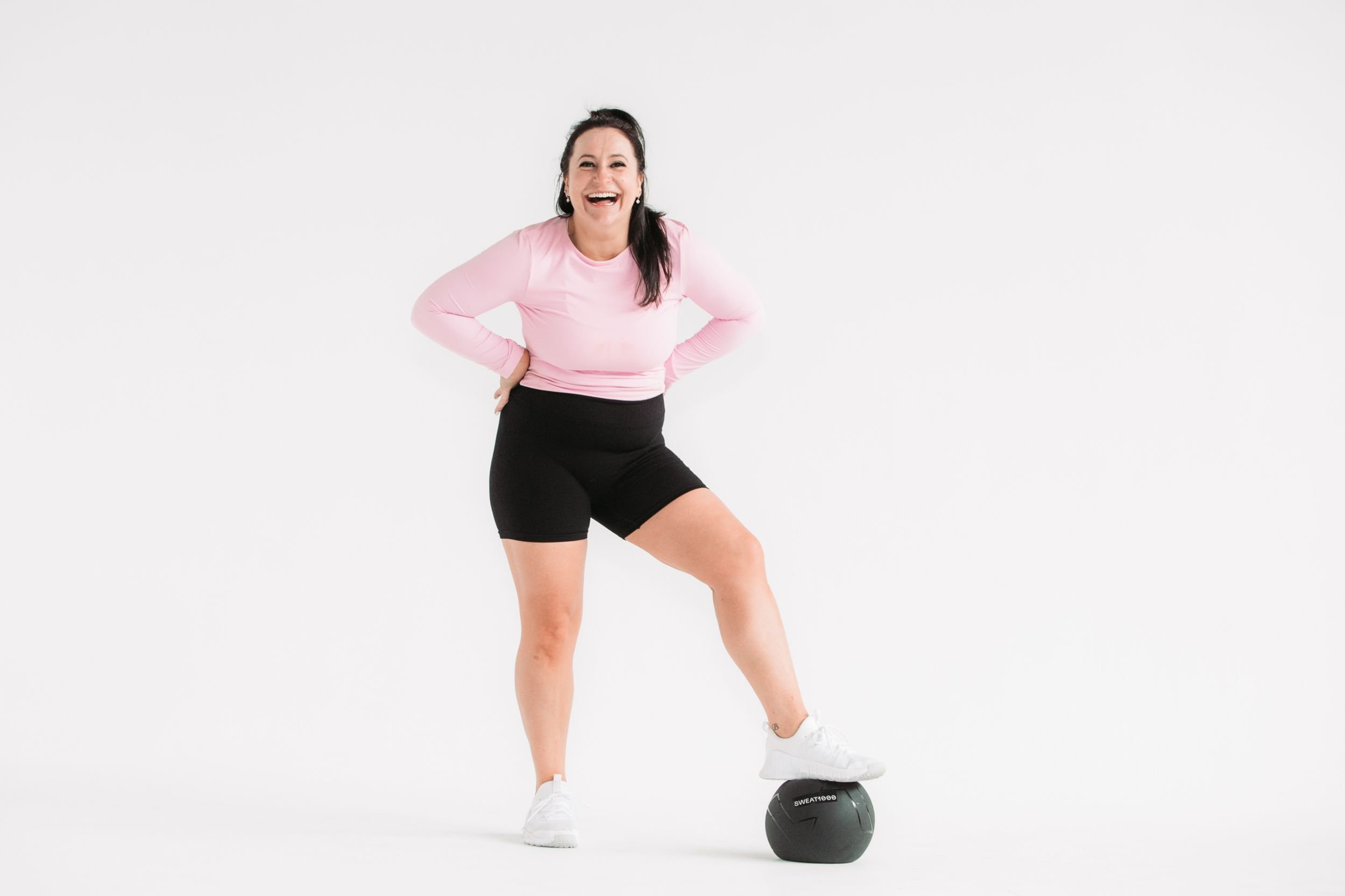 Head coach Kim from MumTribe Fitness, in a pink long-sleeve shirt and black shorts smiling, standing with one foot on a black medicine ball against a plain white background.