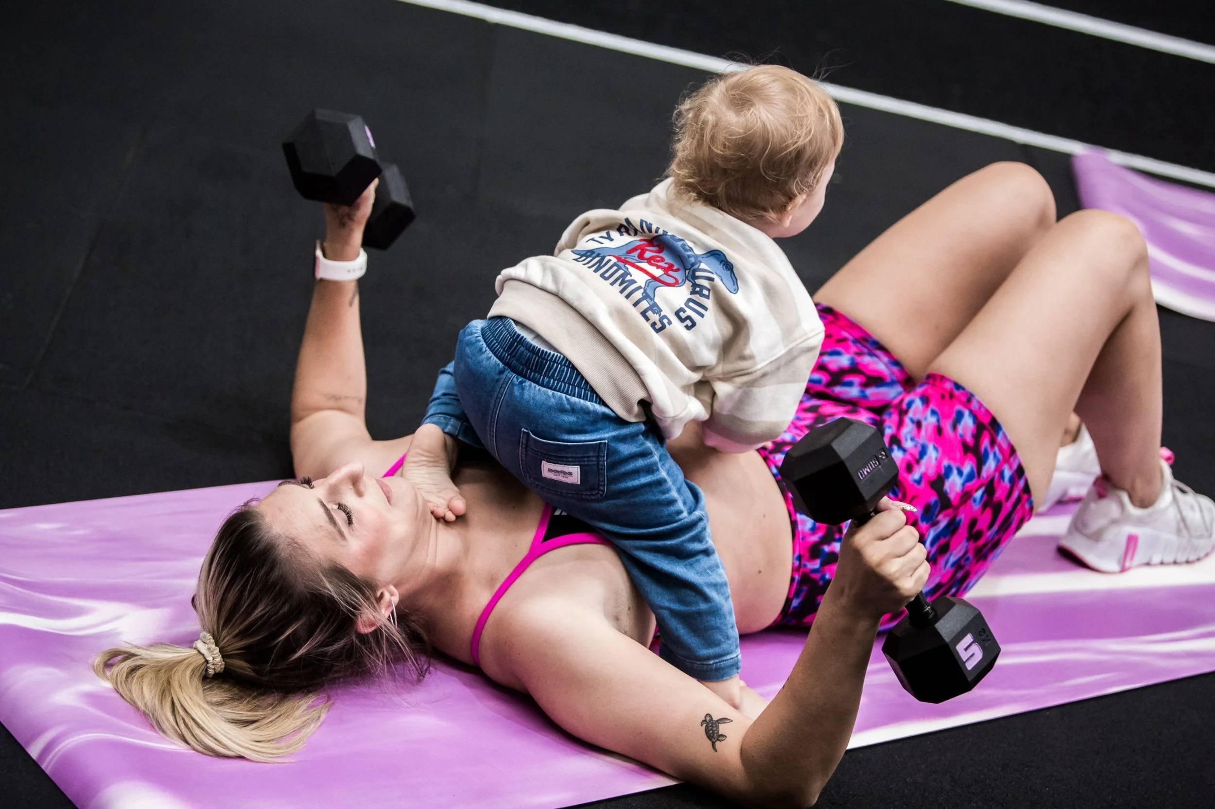 A woman lying on a pink exercise mat lifting dumbbells, with a young child riding on her torso holding smaller dumbbells.