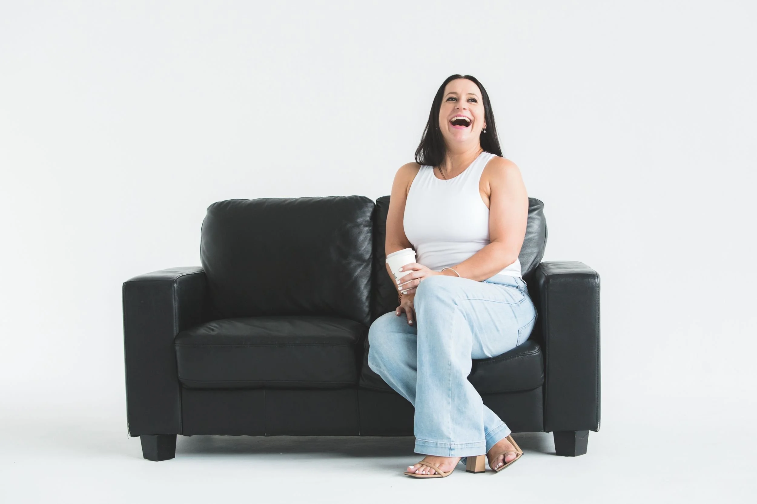 Kim from MumTribe sitting on a black leather sofa, laughing, holding a coffee cup, wearing a white sleeveless top, light blue jeans, and beige high-heeled sandals, against a plain white background.