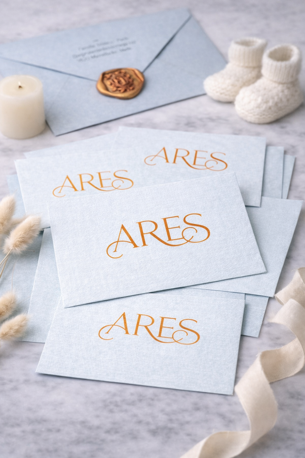 Multiple light blue gift boxes with 'ARES' written in orange on the covers, arranged on a light surface. In the background, there is a light blue envelope with a wax seal, a white candle, and a pair of white baby booties. Additional decorative elements include some dried flowers and a cream-colored ribbon.