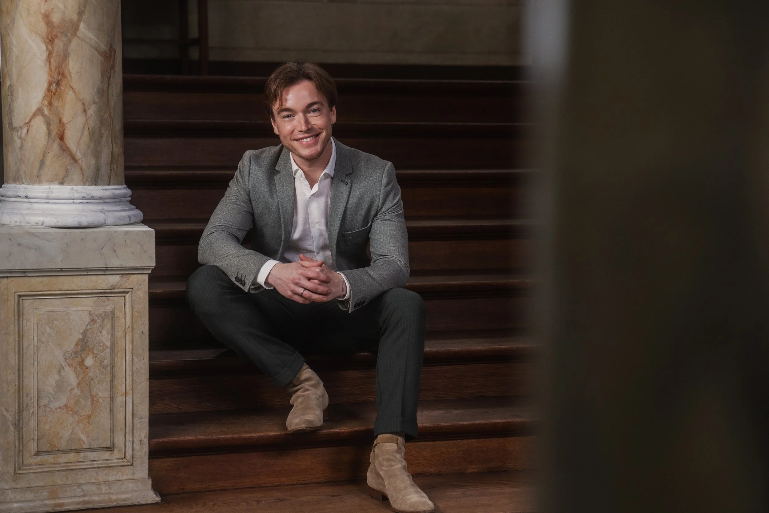 A young man with brown hair, wearing a gray blazer, white shirt, dark pants, and beige boots, sits on wooden stairs inside a building with marble pillars and ornate details, smiling at the camera.
