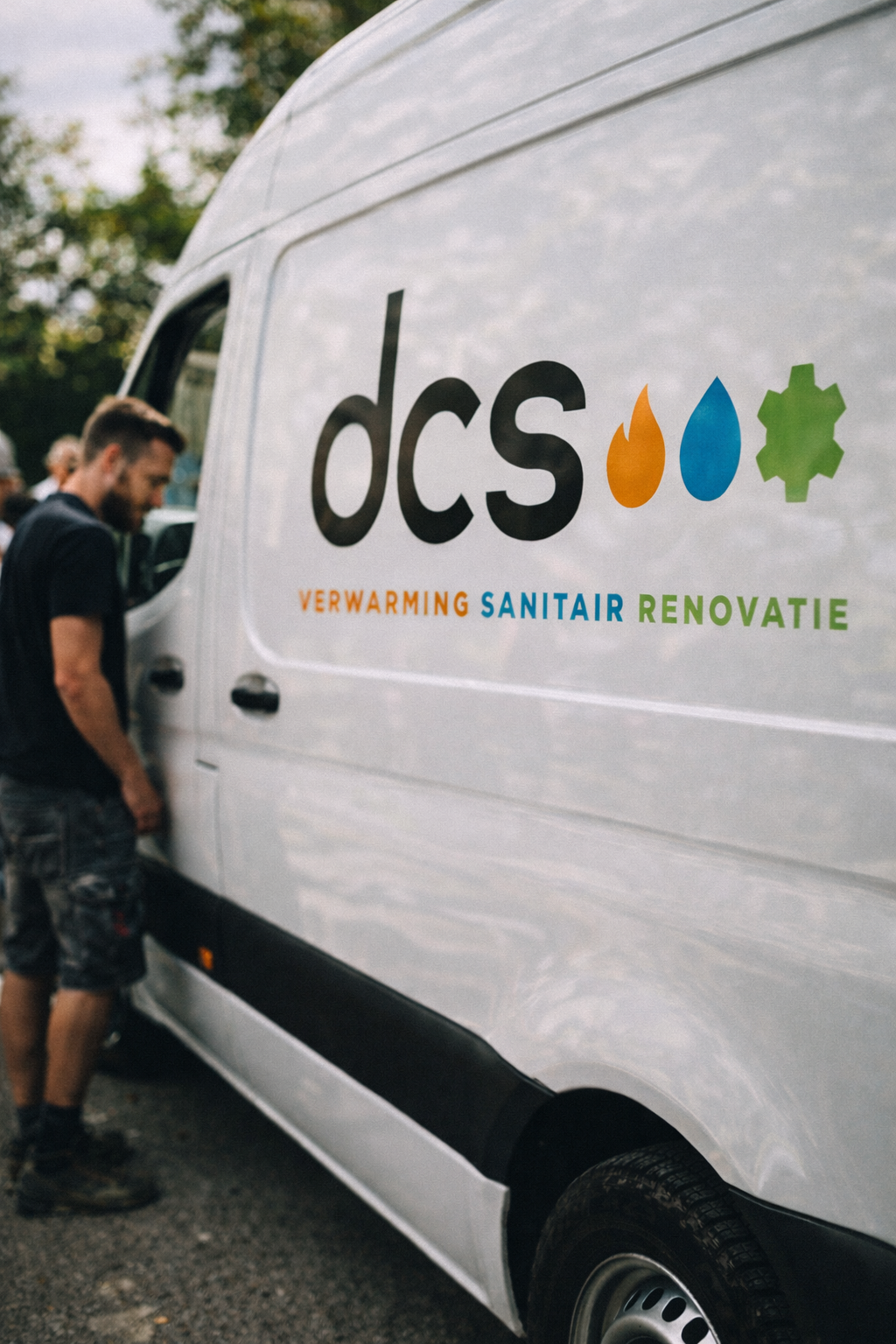 A man standing next to a white van with the logo 'DCS' and symbols of fire, water, and a gear, with words 'Verwarming', 'Sanitair', and 'Renovatie' written below.