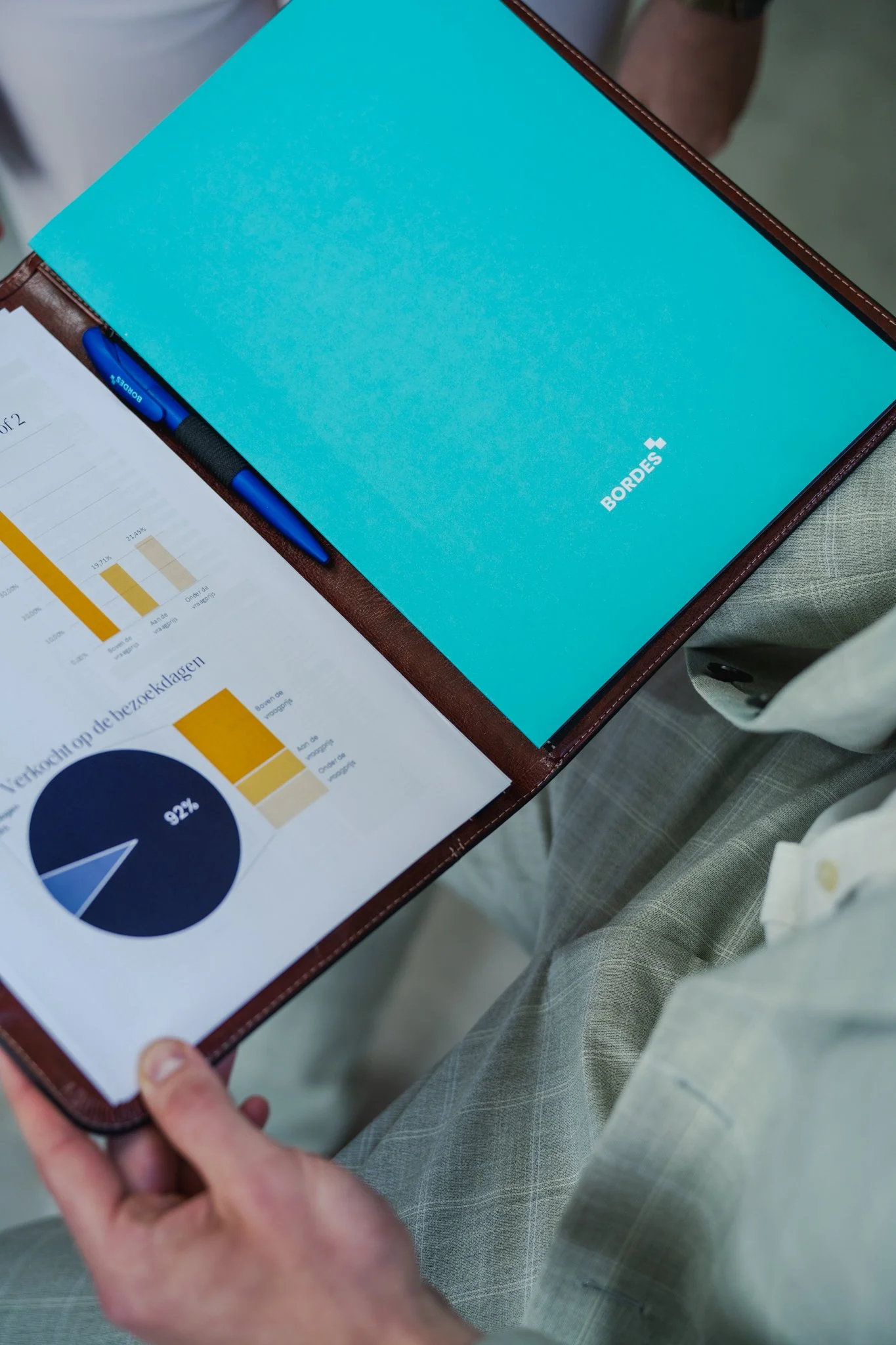 Person holding a leather portfolio with a blue notebook labeled 'BORDES' and a printed report featuring charts.