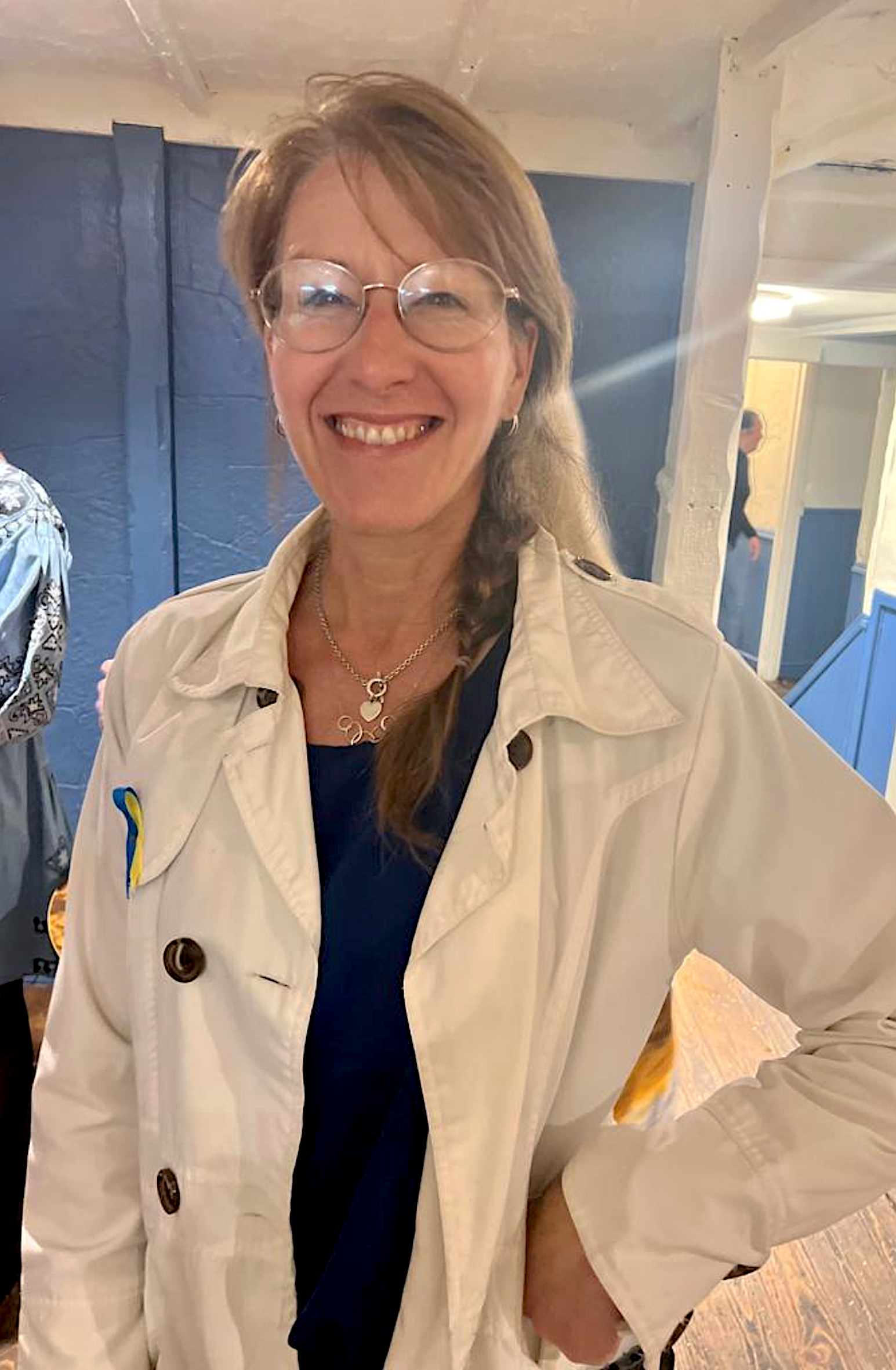 A smiling woman wearing glasses, a beige jacket with a yellow and blue ribbon pin, and a necklace, standing indoors in a well-lit space.