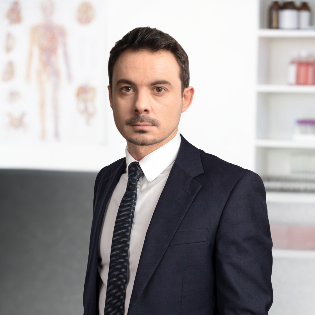 A man in a dark suit and tie standing in an office with medical posters and shelves in the background.
