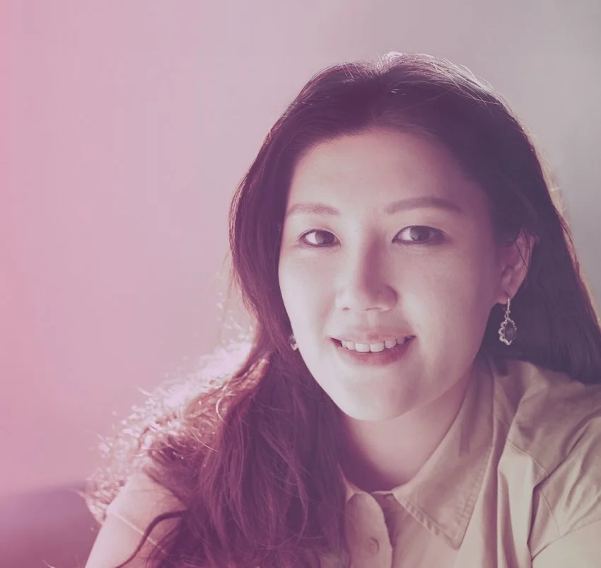 A young woman with dark hair, wearing earrings and a beige shirt, smiling softly in a softly lit room.