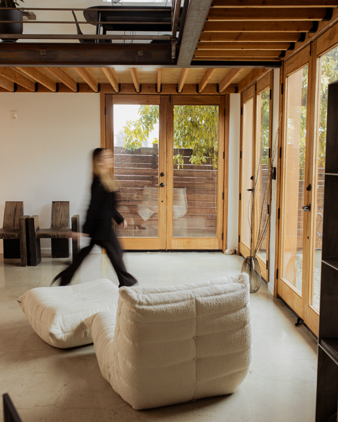 Modern living room with wooden doors and large windows, cream-colored armchair, small seating area, and a person walking.