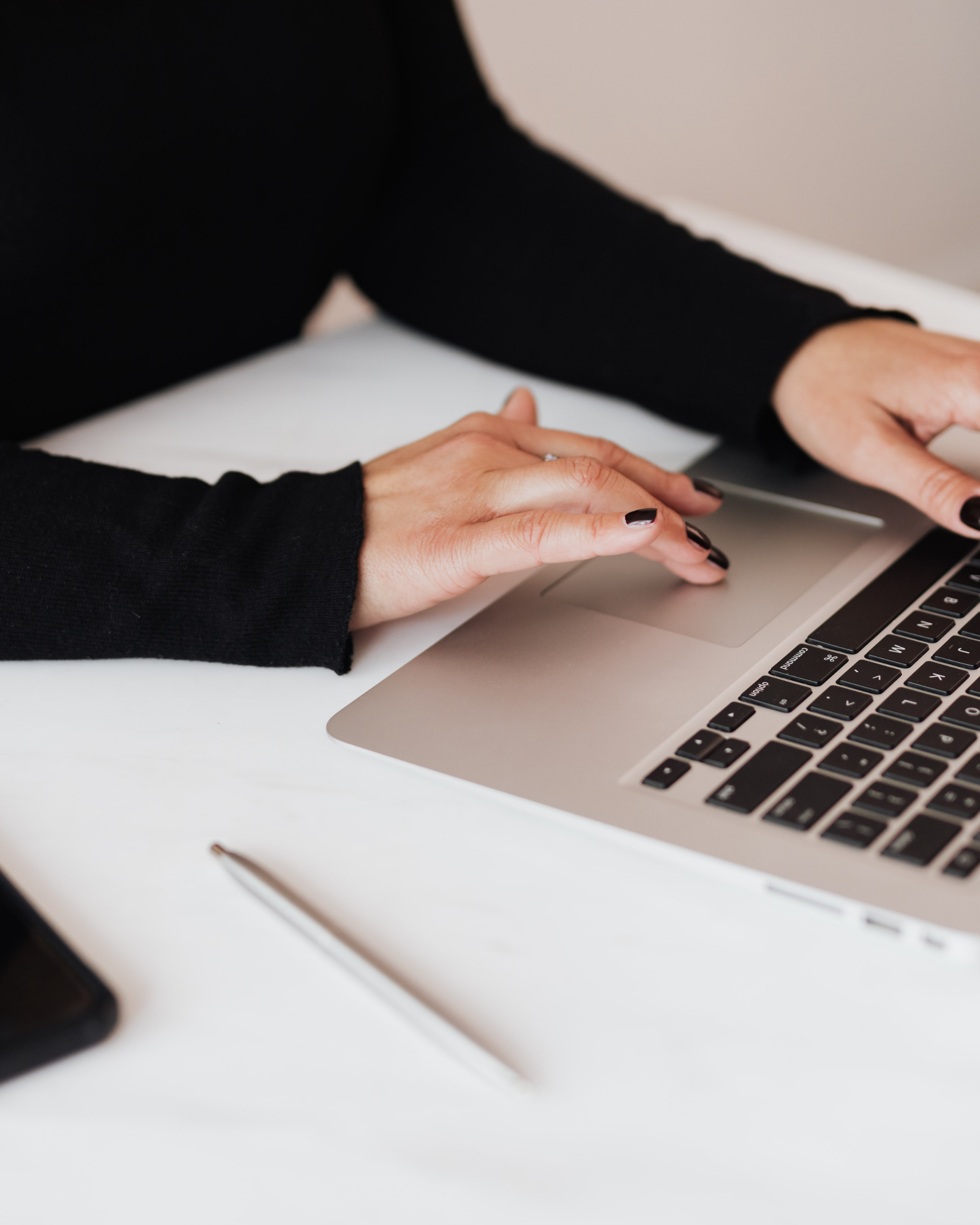 Close-up of two hands typing on a silver laptop keyboard on a white desk, with a pen lying nearby.