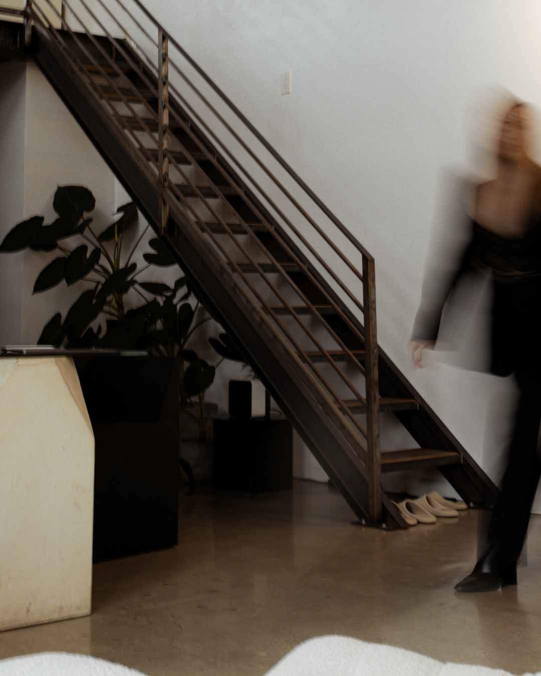 Indoor scene with an office staircase and a blurred person walking by, with shoes under the stairs, a large plant, and a desk on the left.