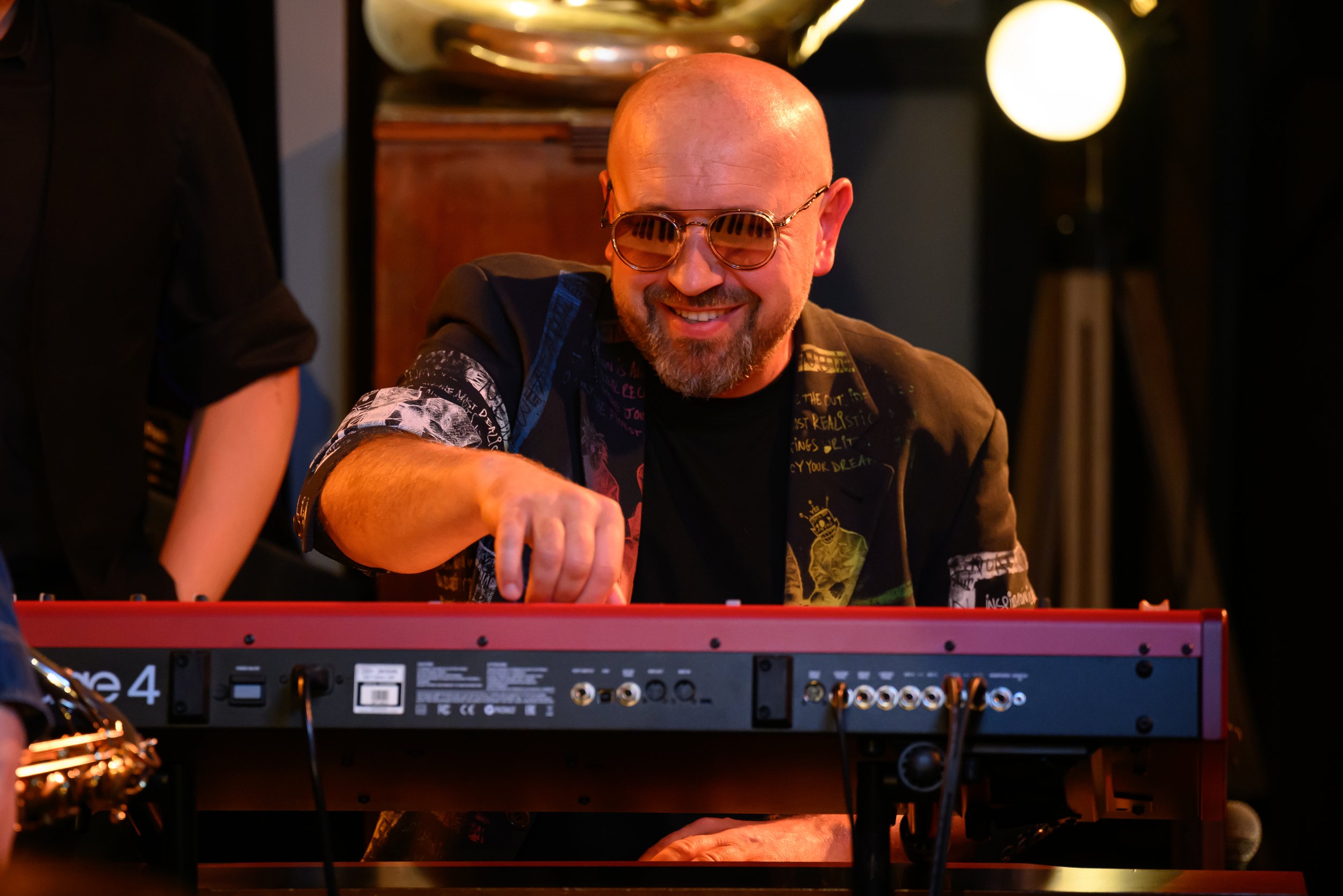A man wearing sunglasses and a colorful jacket playing a red keyboard on stage.
