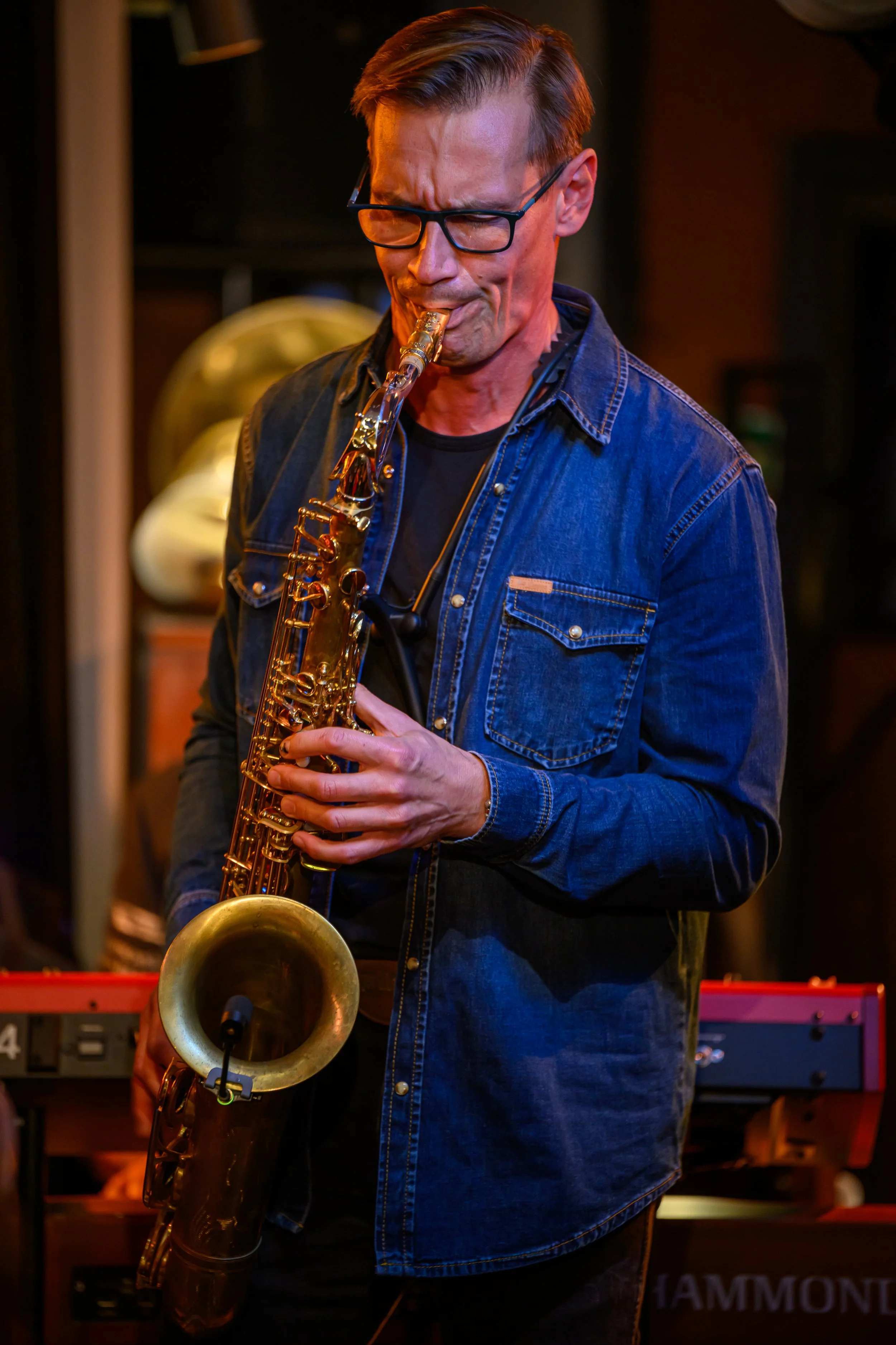 A man wearing glasses and a denim jacket playing a saxophone indoors.