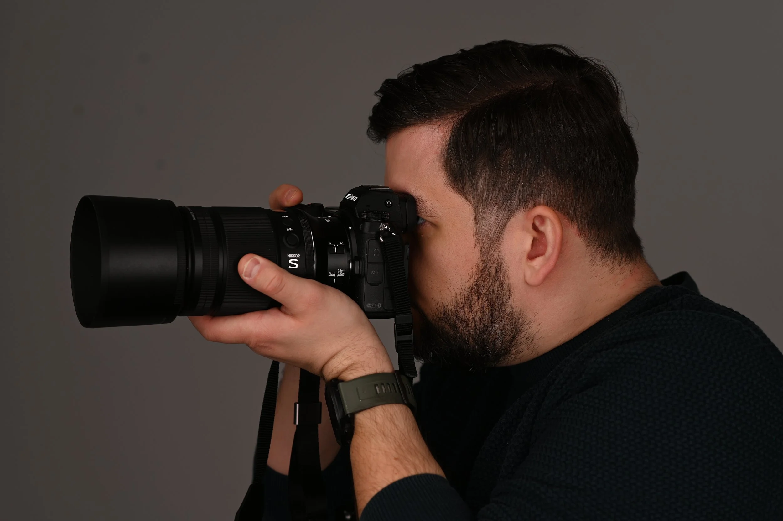A man with dark hair and a beard is taking a photograph using a professional camera with a large telephoto lens against a plain gray background.