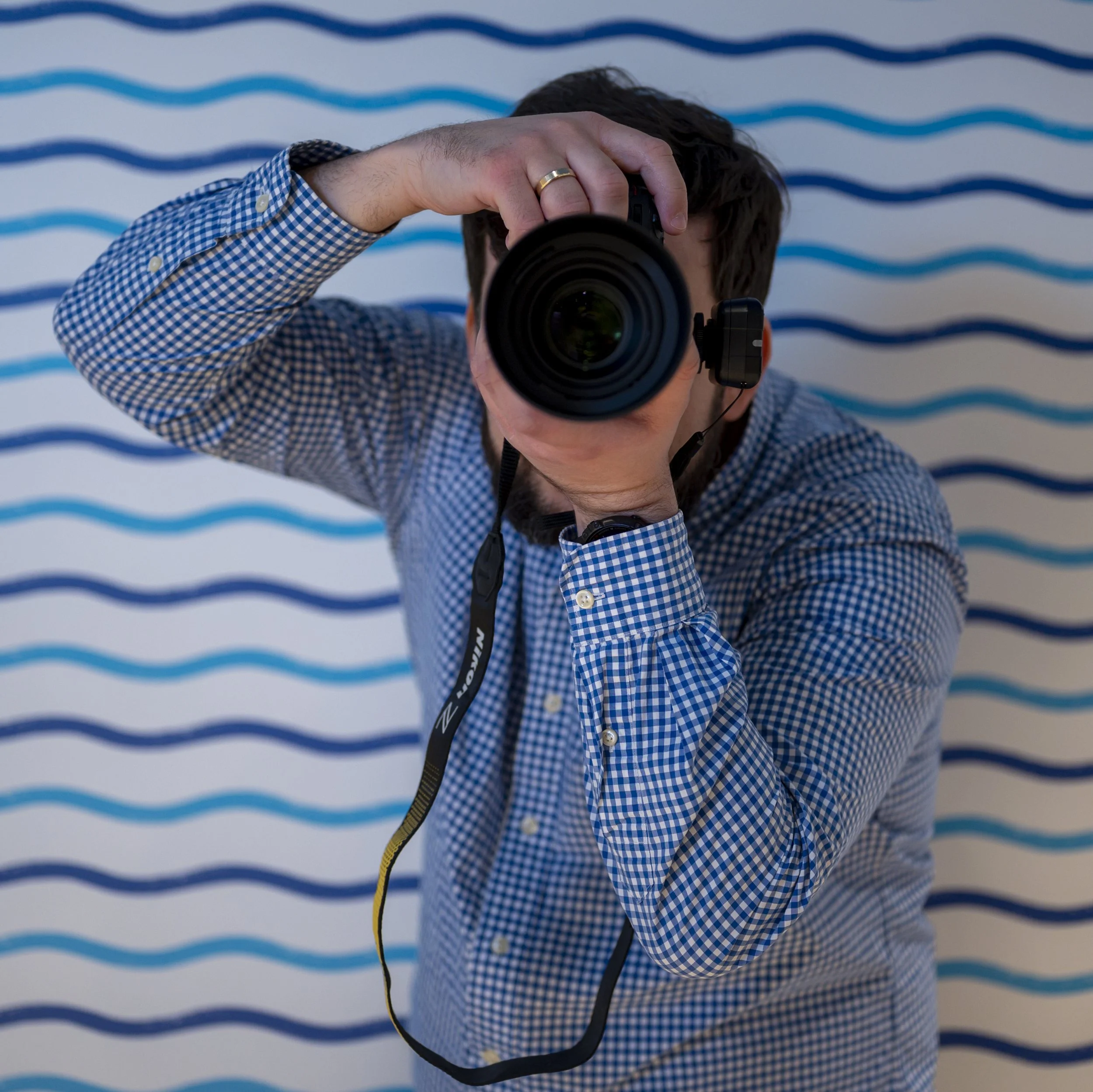 A man wearing a blue checkered shirt taking a photo with a camera, standing in front of a white wall with blue wavy lines.