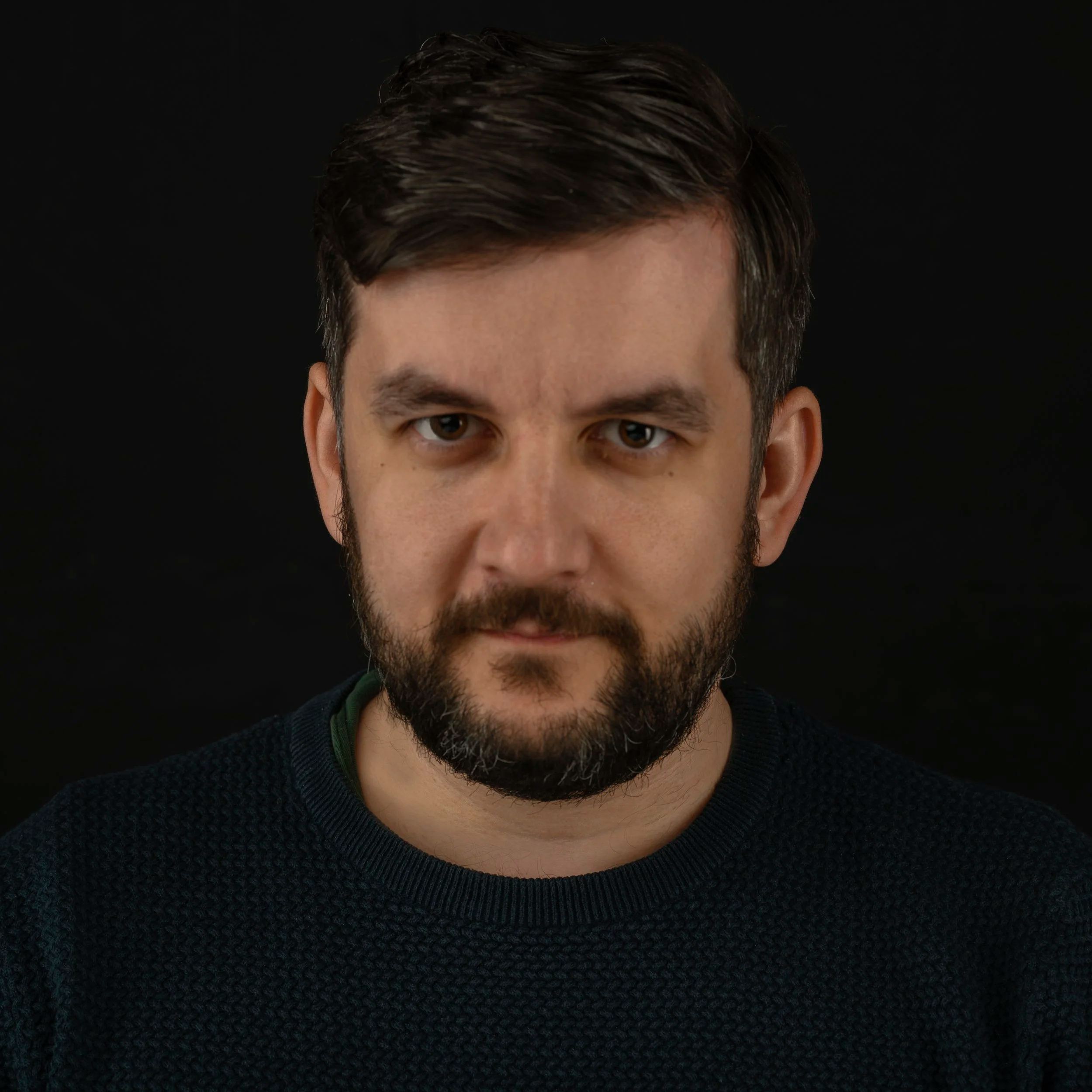 Close-up of a man with dark hair and a beard against a black background.
